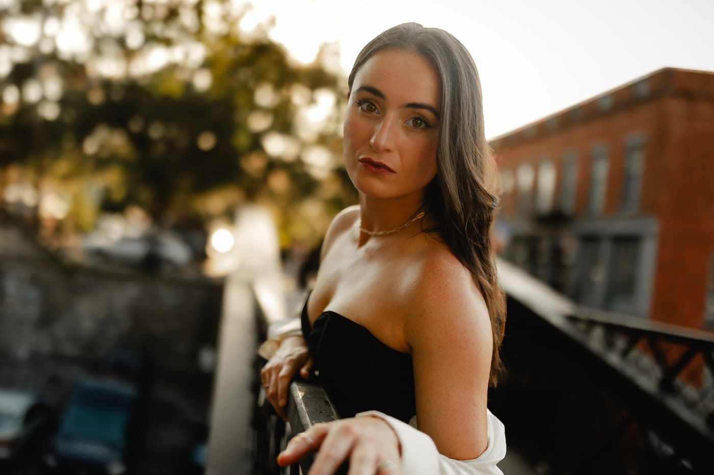 A young brunette woman in a black strapless top poses outdoors on a urban balcony at golden hour.