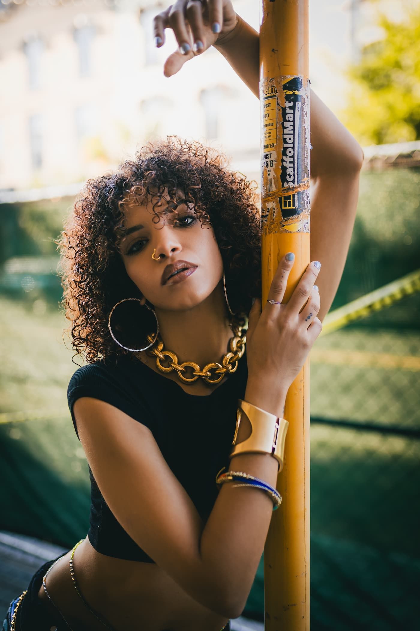 Stylish woman with curly hair posing with gold chain necklace and hoop earrings by yellow pole.