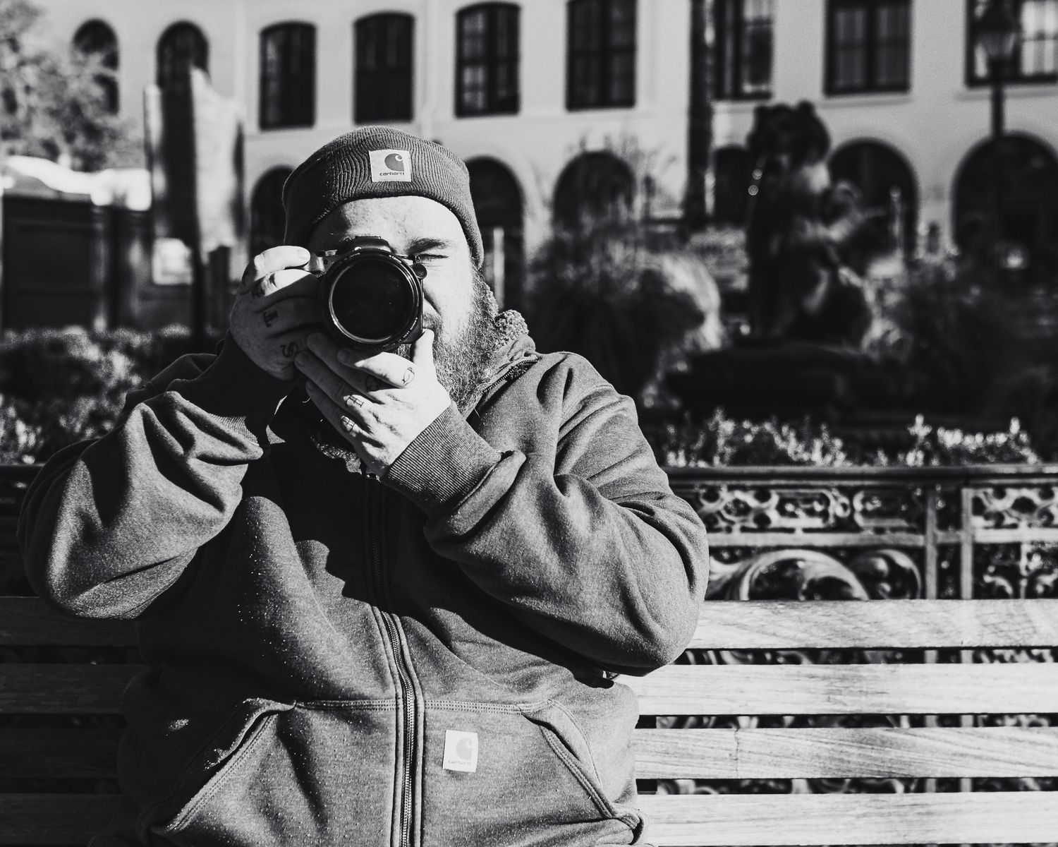 Black and white photo of a bearded man in a Carhartt hoodie and beanie photographing with a DSLR camera on a park bench.