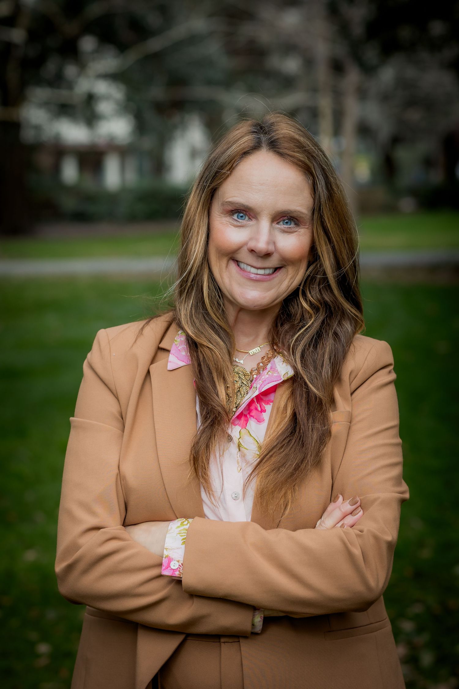 Professional woman in tan blazer with arms crossed, smiling outdoors in a green park setting.