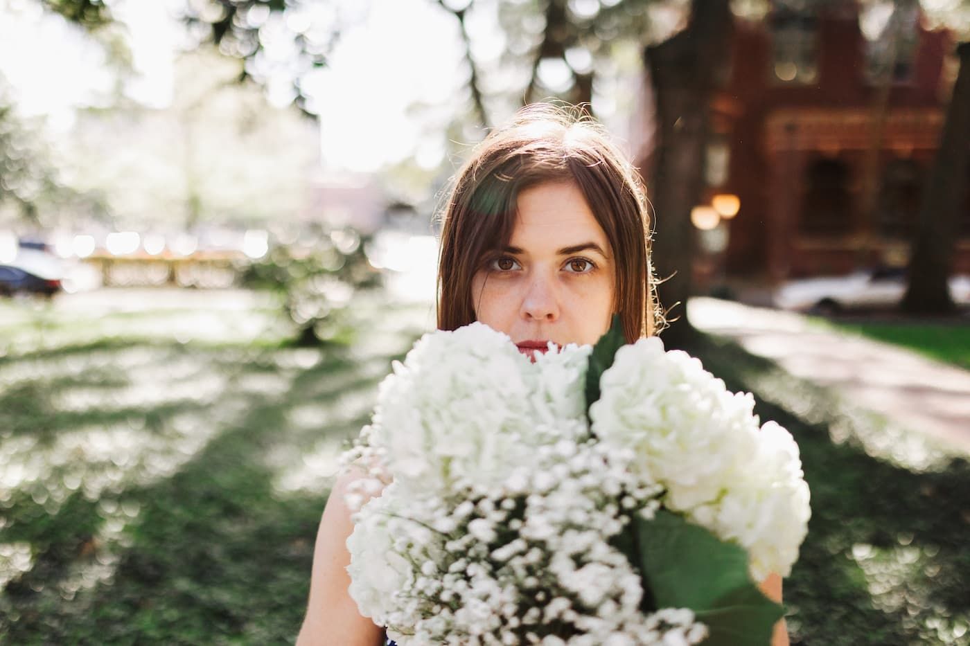 Young woman holding large bouquet of white hydrangeas outdoors in sunlit park setting.