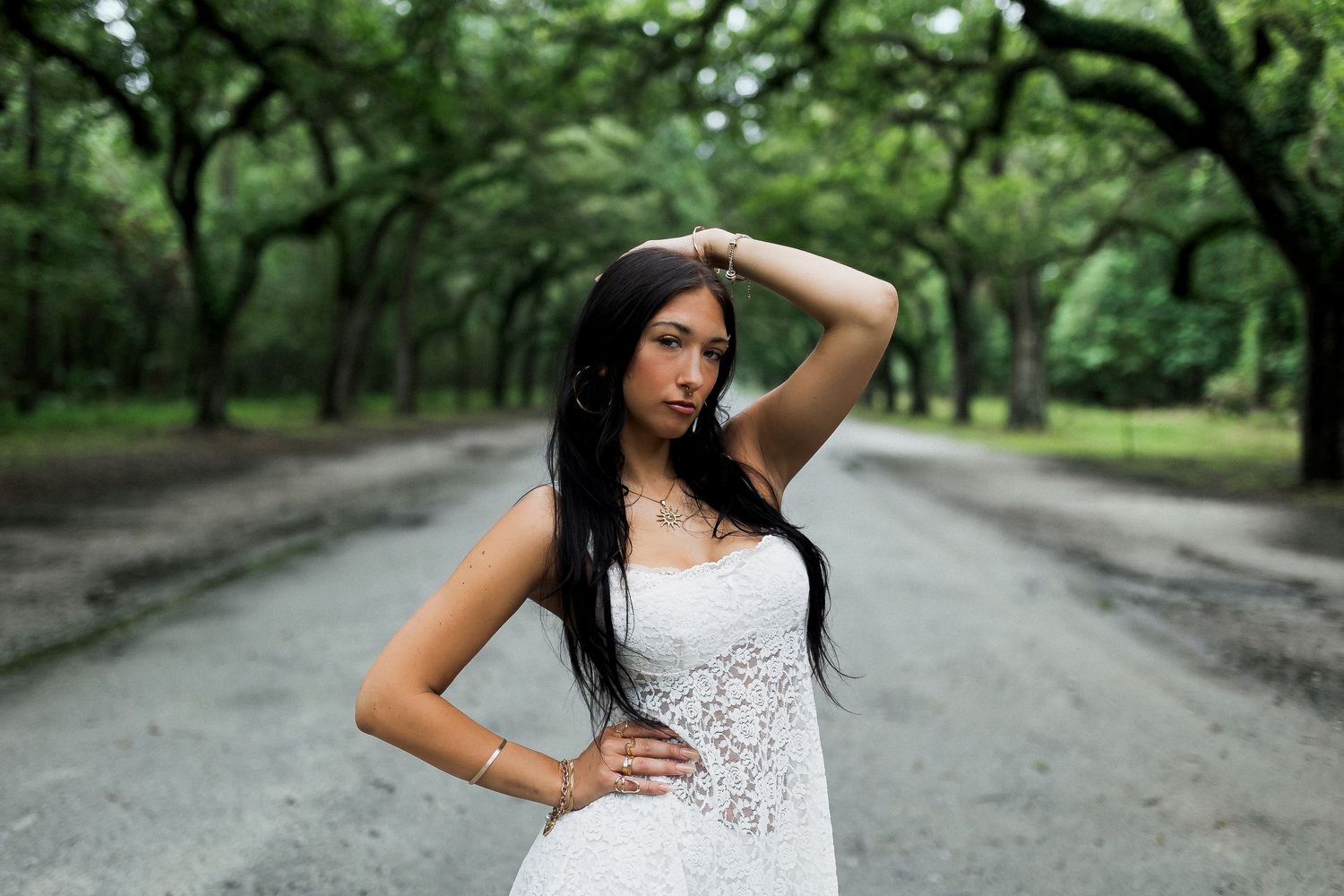 Young woman in white lace dress posing on a tree-lined road surrounded by lush green trees.