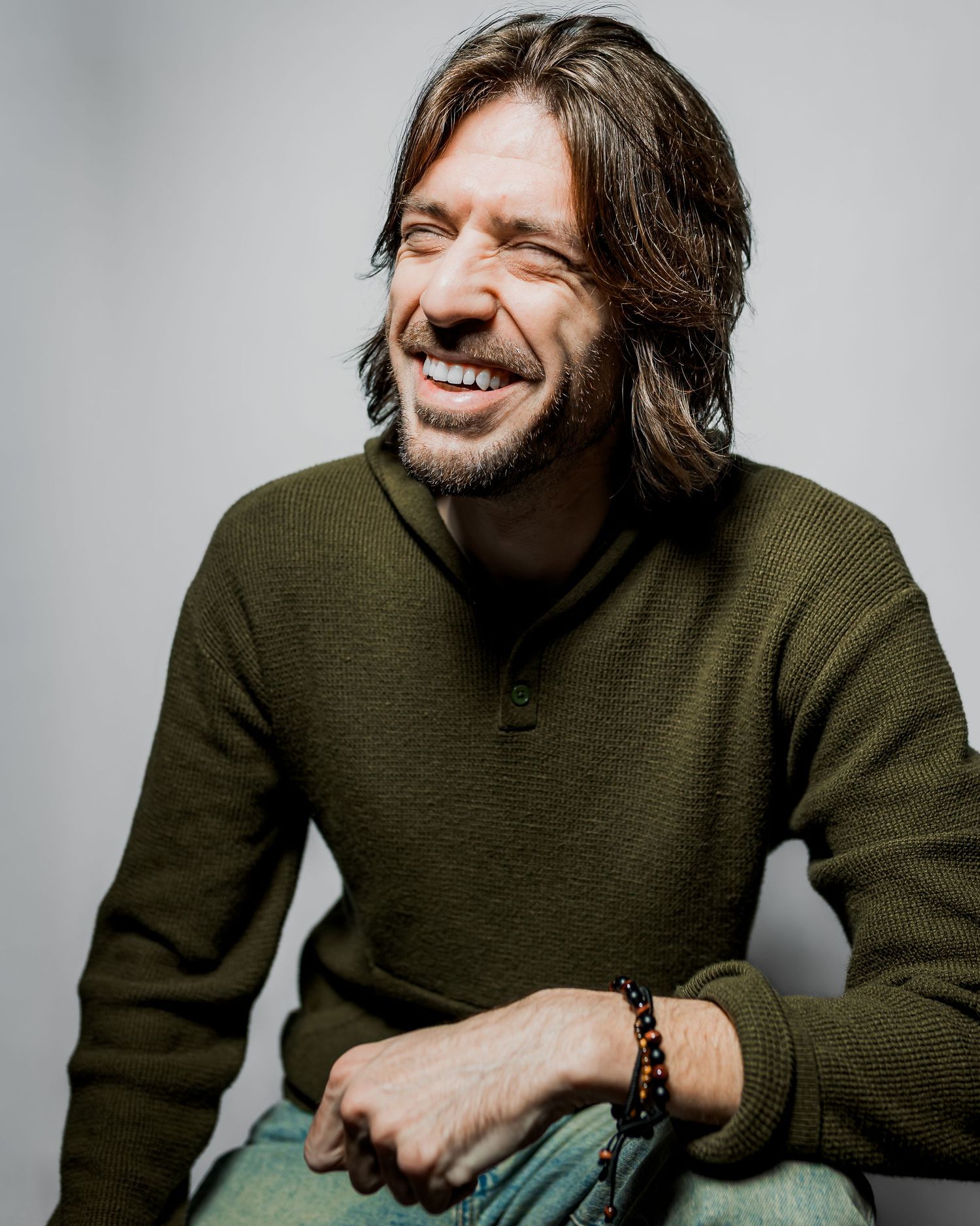 Smiling man with long brown hair wearing a green sweater and beaded bracelet, laughing in a studio portrait.