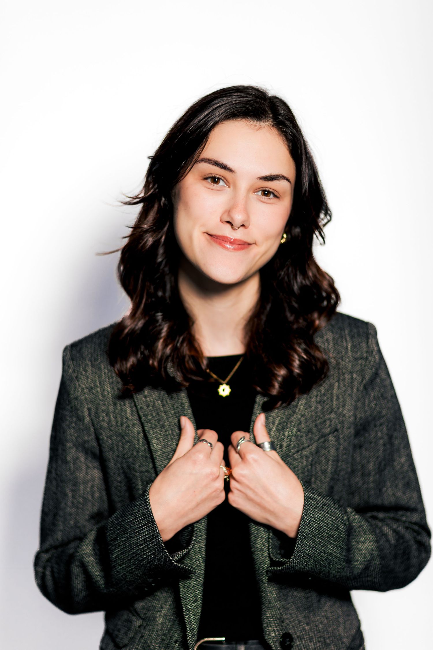 Young woman in gray blazer smiling confidently with hands clasped, wearing gold necklace, against white background.