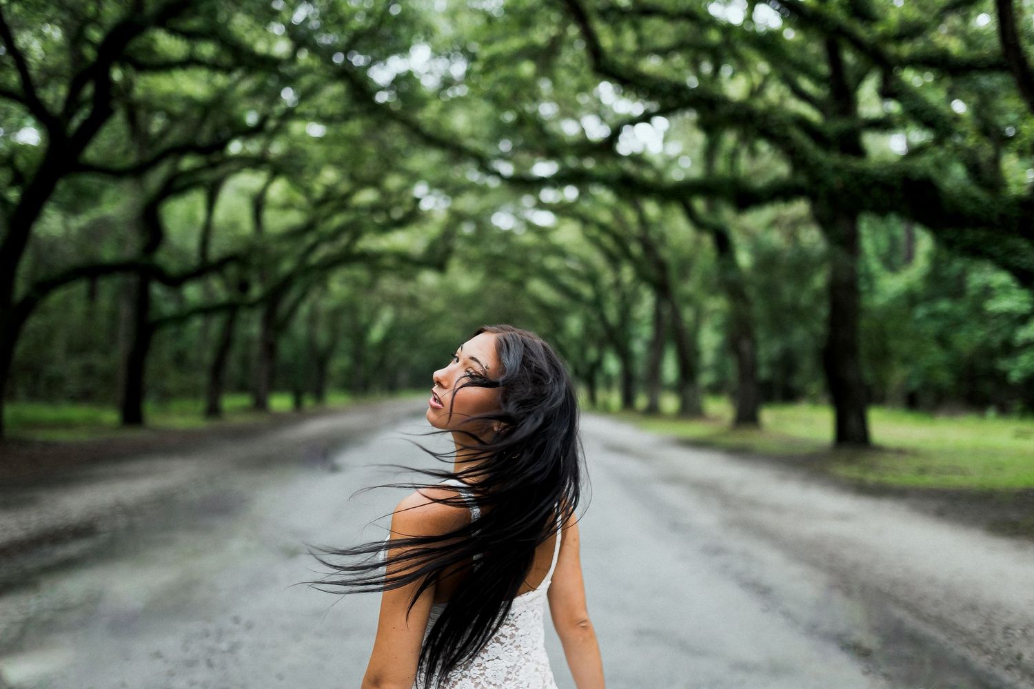 Woman tossing long dark hair on a tree-lined road surrounded by lush green canopy.