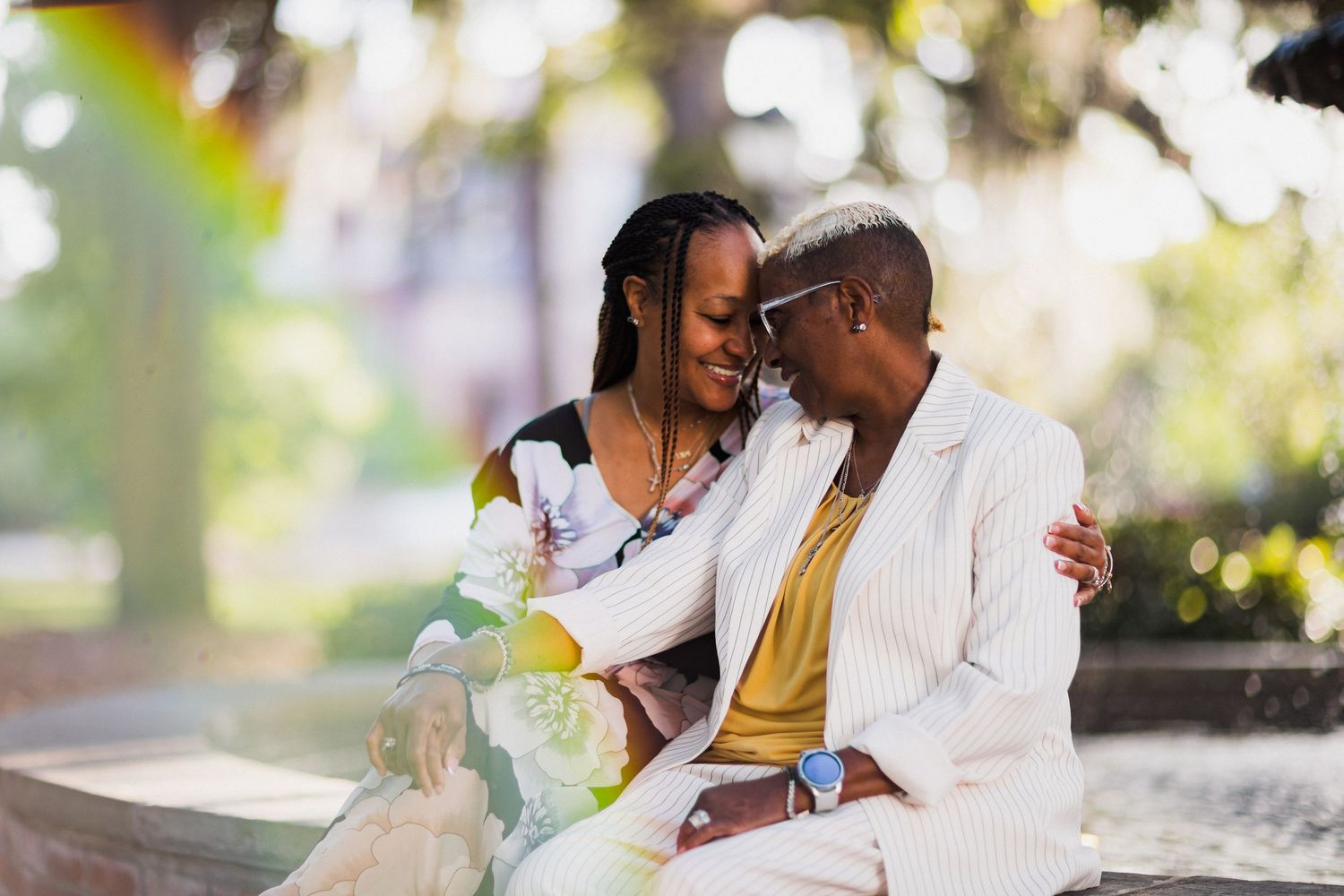 Two women sharing a tender moment, smiling and embracing outdoors in a sunlit park setting.