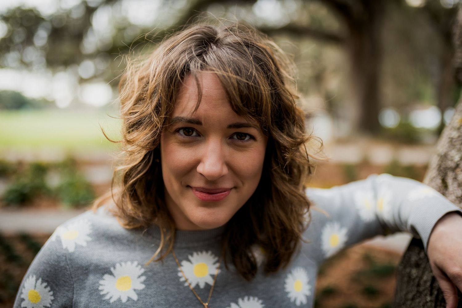 Young woman with curly brown hair wearing a daisy print sweater, smiling outdoors near a tree.