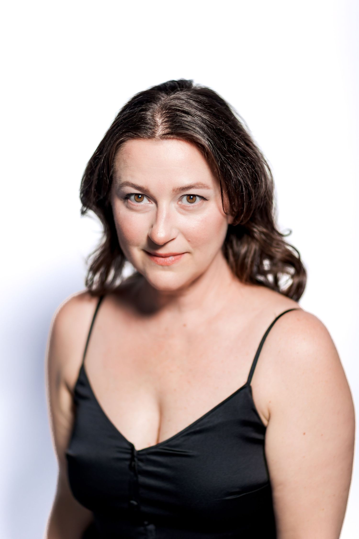 A woman with dark wavy hair wearing a black spaghetti strap top poses against a white background.