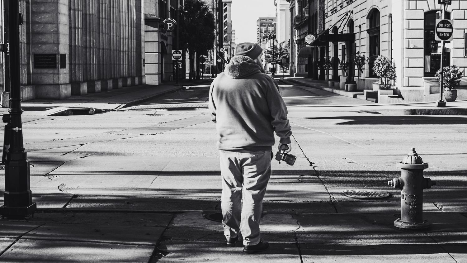 Black and white photo of an elderly man walking with a cane on a city sidewalk past a fire hydrant.