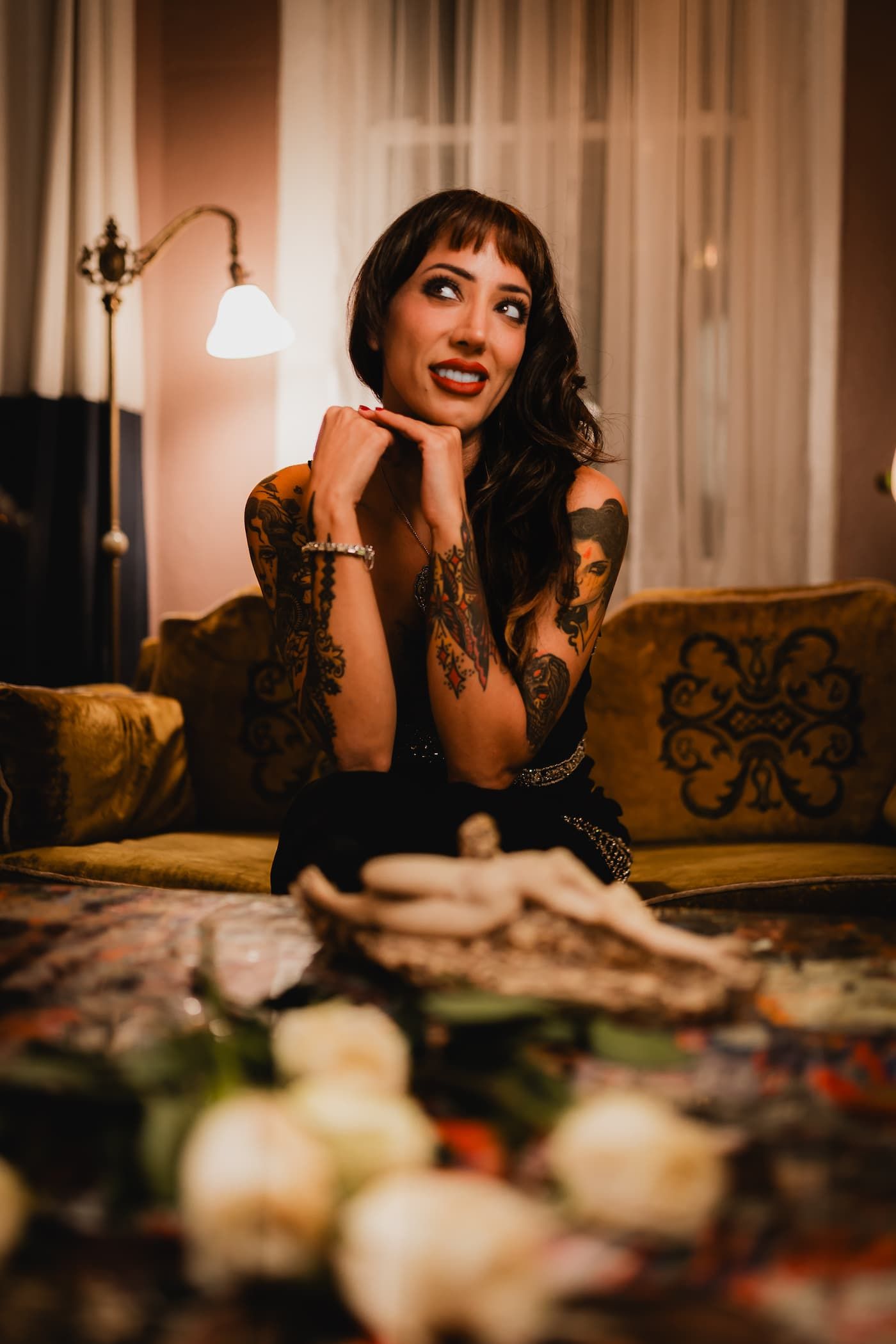 Tattooed woman with dark hair sits on a vintage sofa, smiling dreamily, with white roses in the foreground.