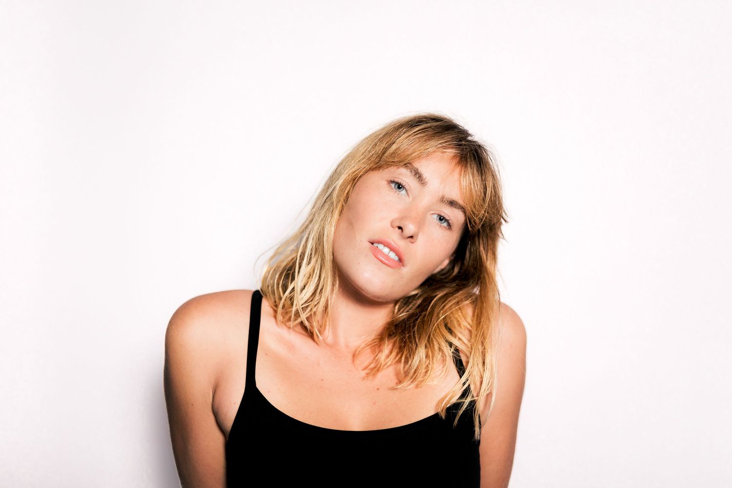 Young blonde woman in black tank top posing against white background with head tilted.