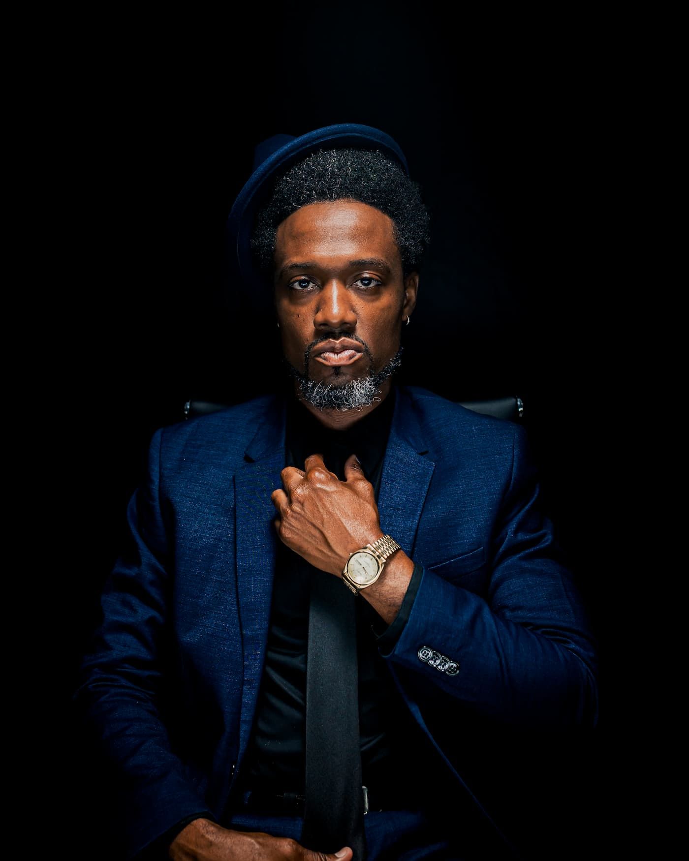 A well-dressed Black man in a blue suit and hat poses confidently with hands clasped, wearing a gold watch.