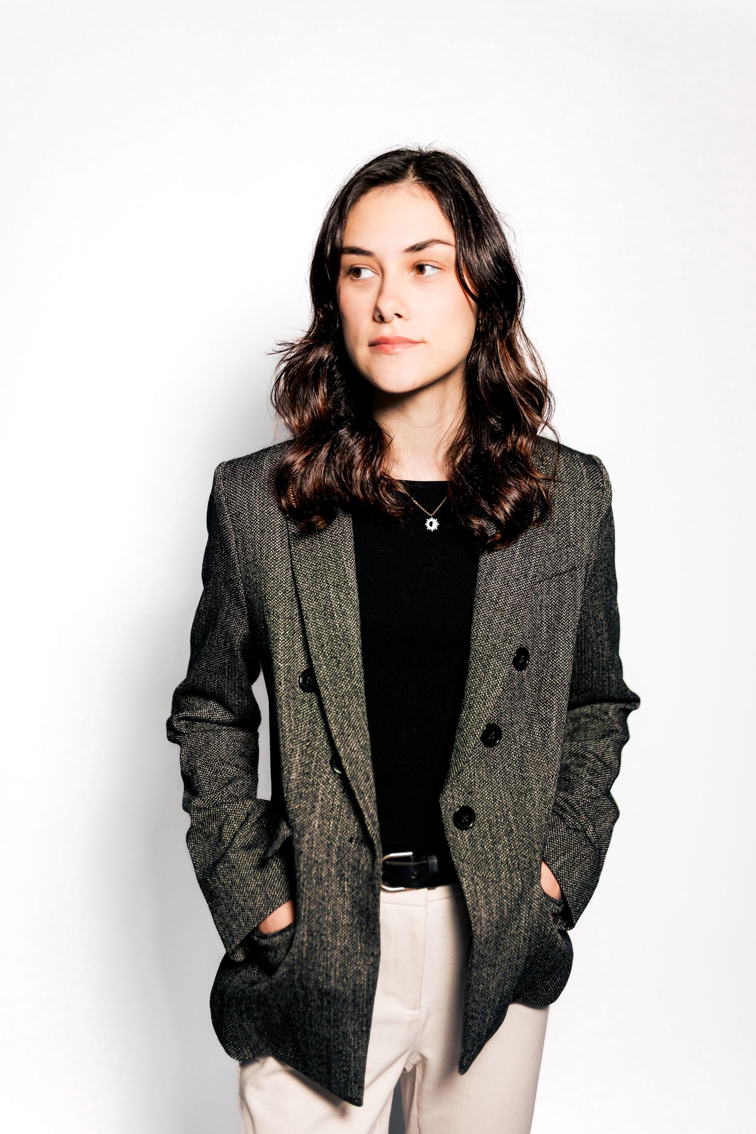 Young woman in a gray herringbone blazer over black top, hands in pockets, posing against white background.