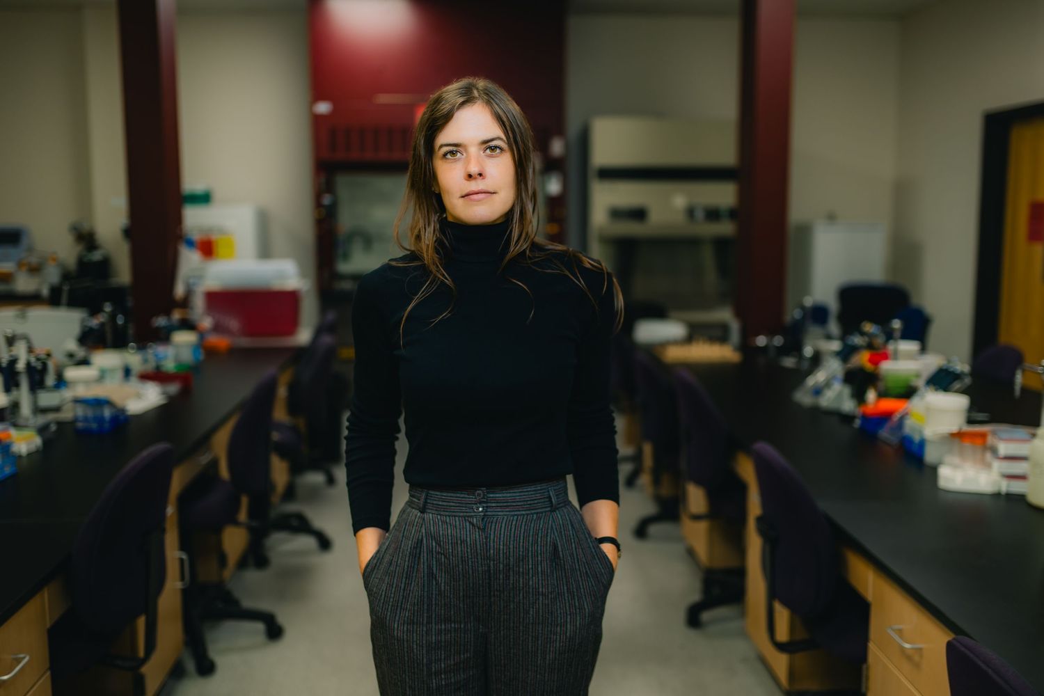 Young woman in black turtleneck and gray pants stands confidently in a modern science laboratory.
