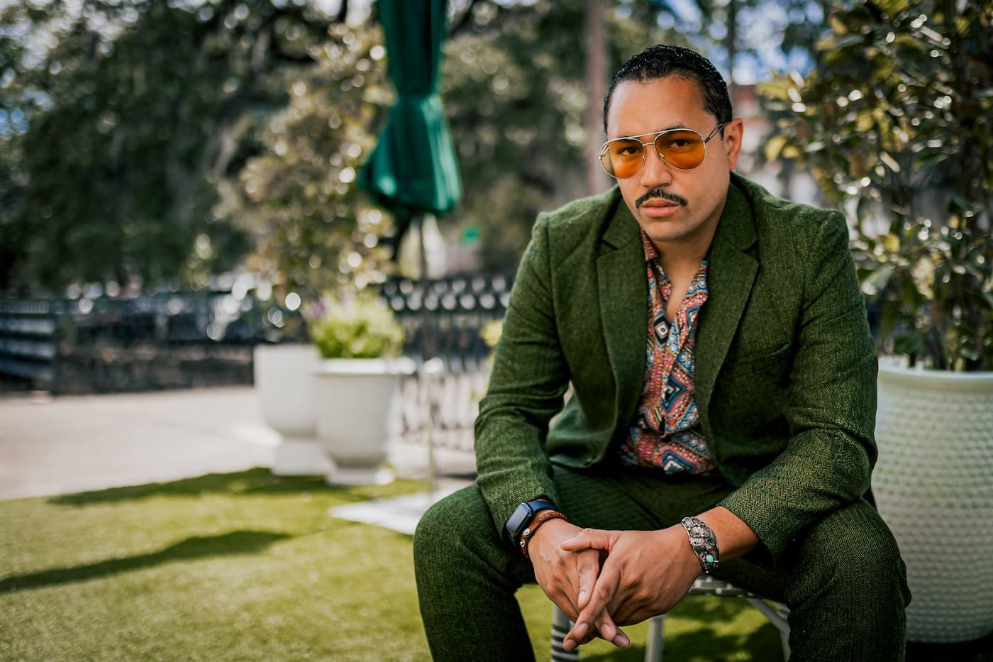 Stylish man in green suit and orange sunglasses sitting outdoors in a garden setting.