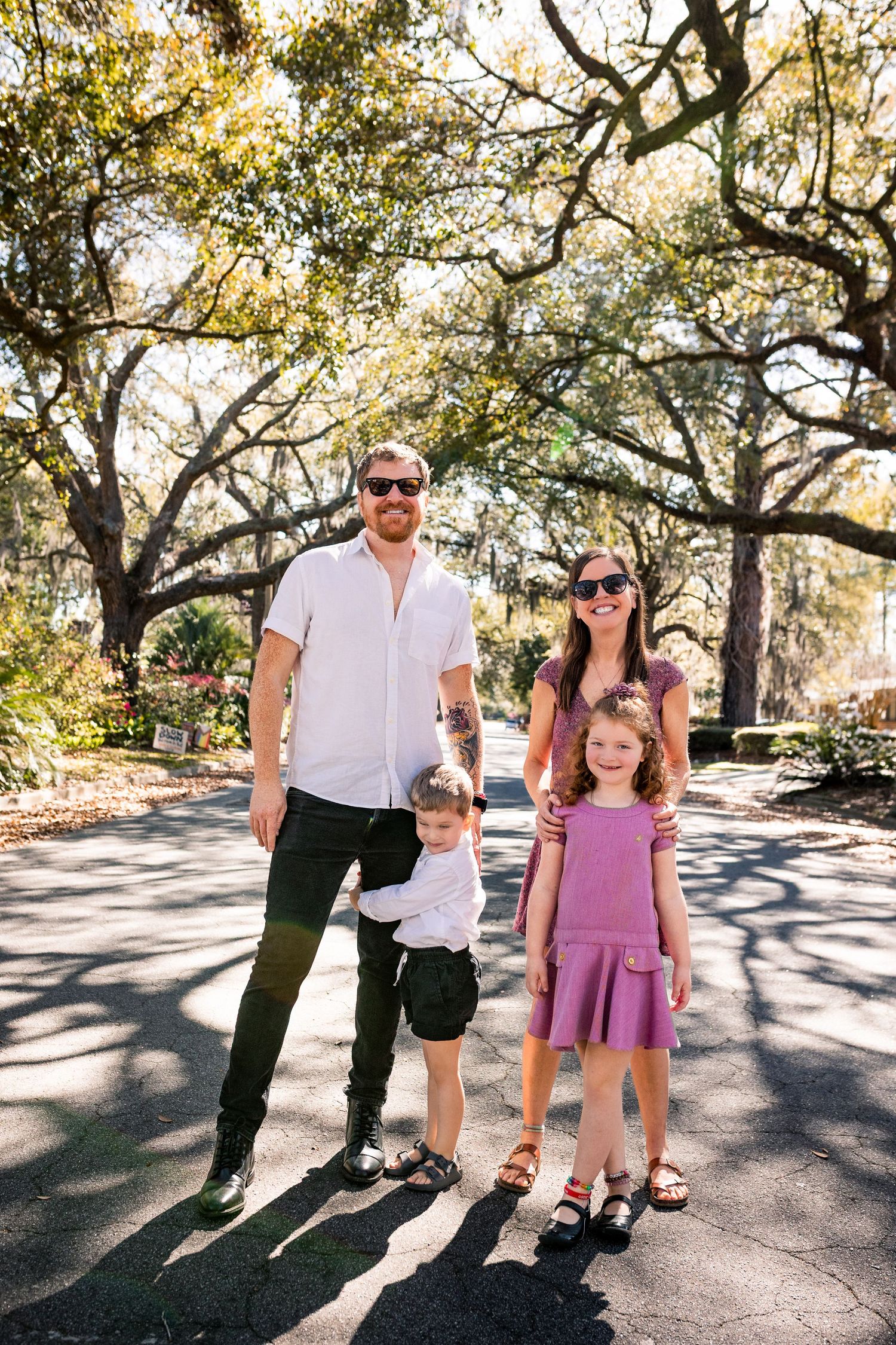 Family of four posing outdoors under oak trees on a sunny day, parents in sunglasses with two young children in purple outfits.