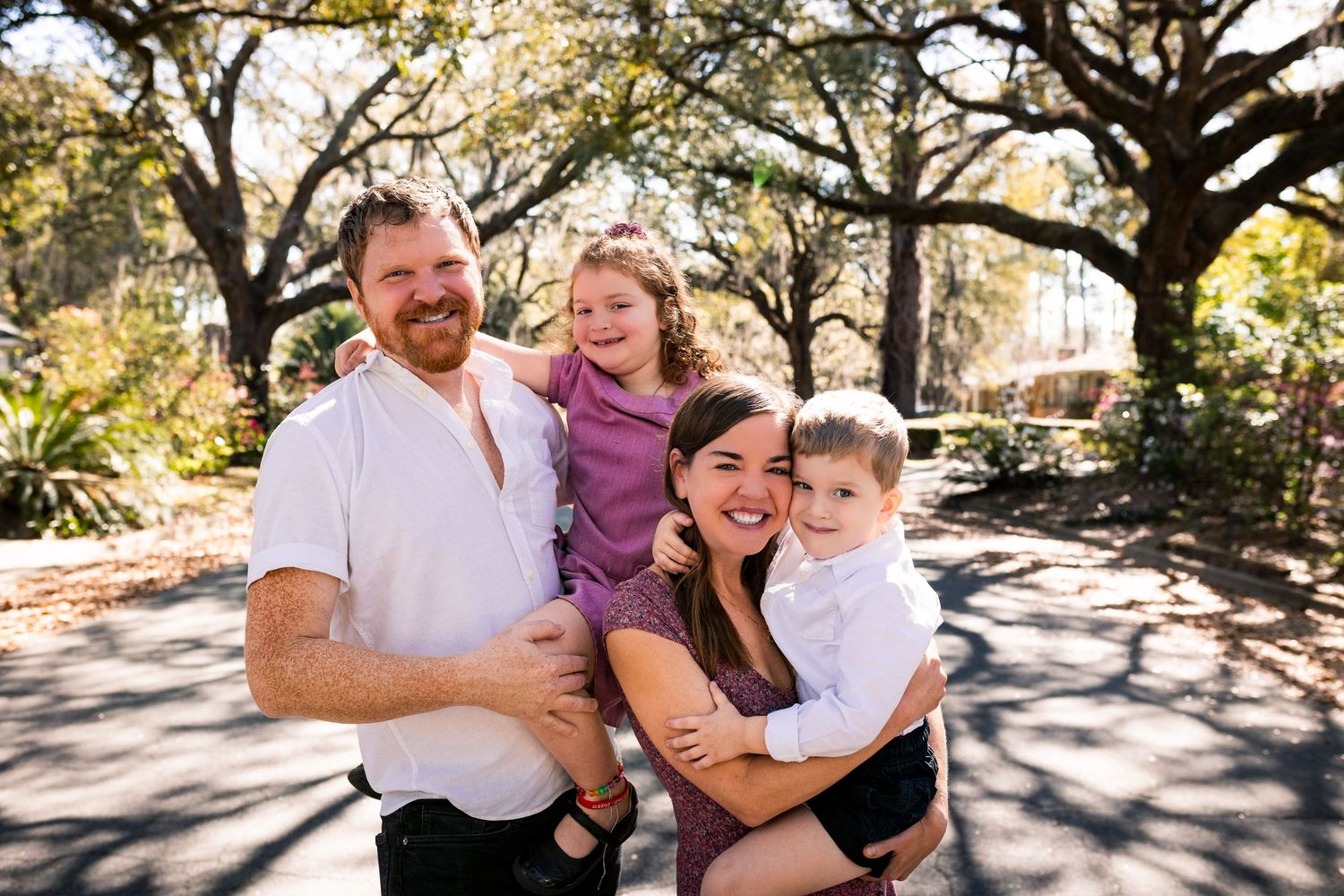 Happy family of four poses outdoors under oak trees, parents holding two young children, smiling in warm natural light.