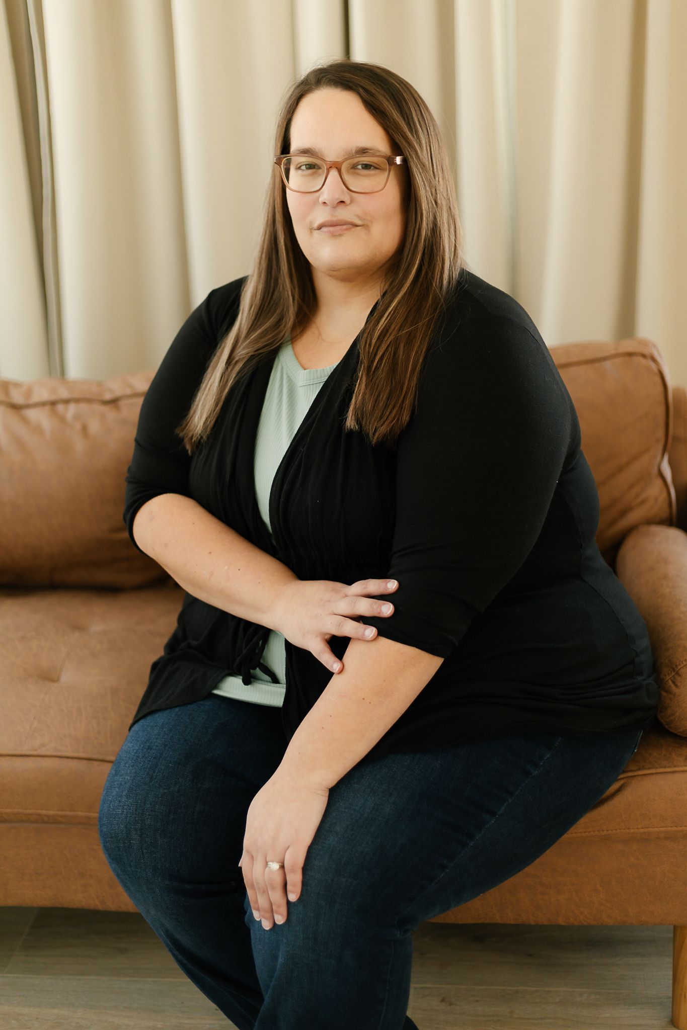 A woman in a black cardigan and mint green top smiles warmly while sitting on a tan leather couch indoor studio, elkton SD