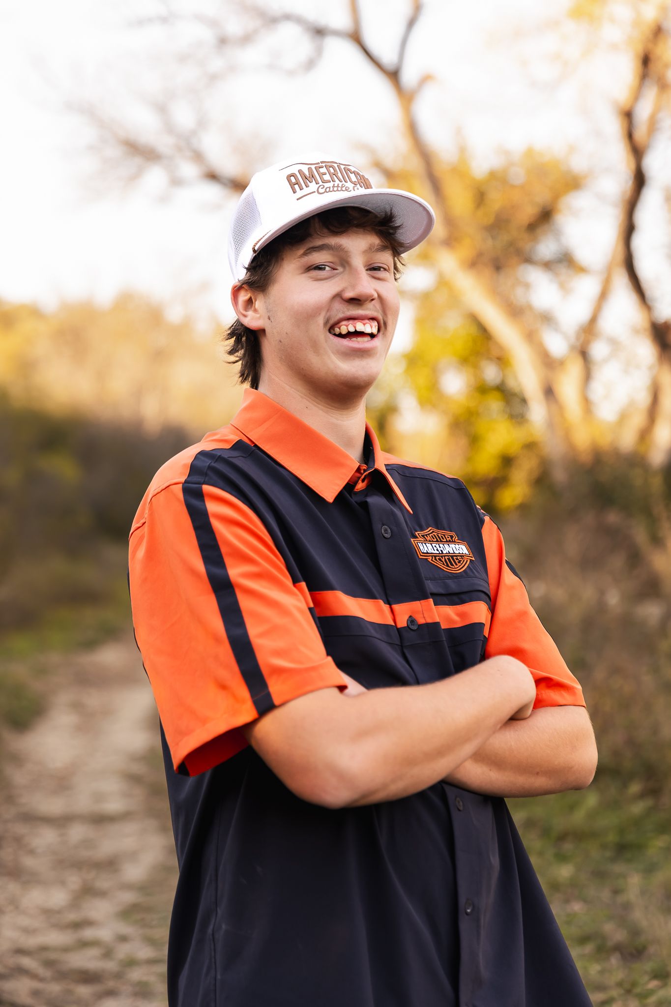 Senior in blue jeans and orange harley vest stands on dirt path during golden hour in Lake Benton, MN