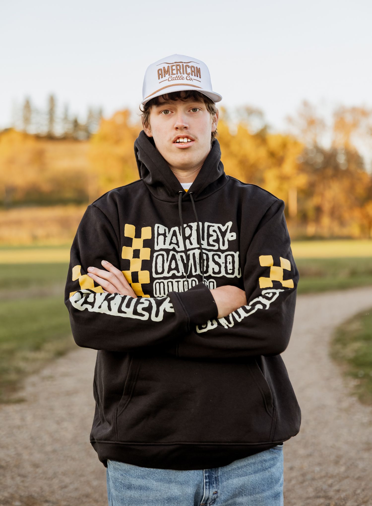 Senior wearing a black Harley-Davidson hoodie and baseball cap stands on a dirt path during autumn season in lake benton, MN