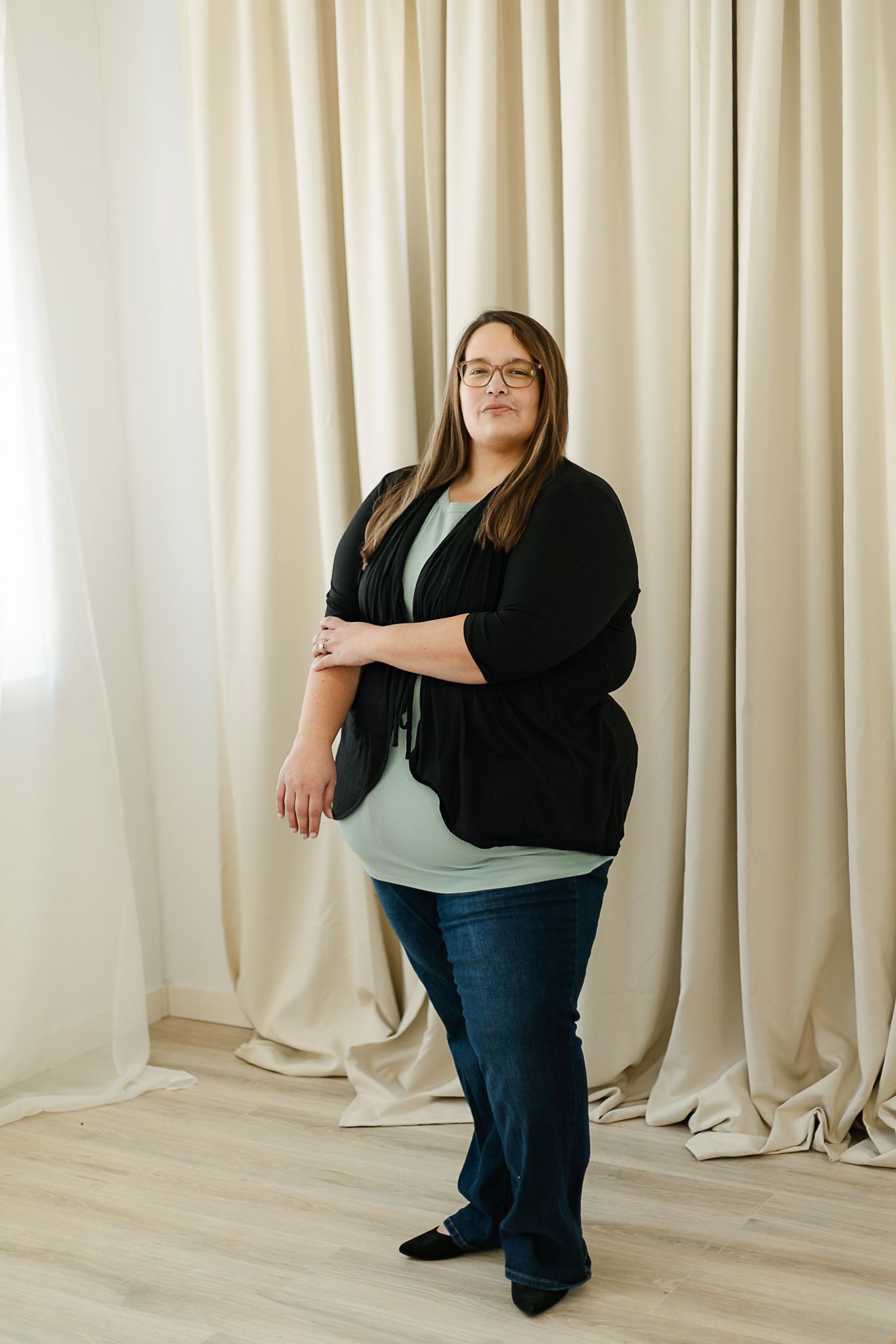 A woman in a black cardigan and mint green top smiles warmly while posing against a cream-colored curtain backdrop indoor studio, elkton SD