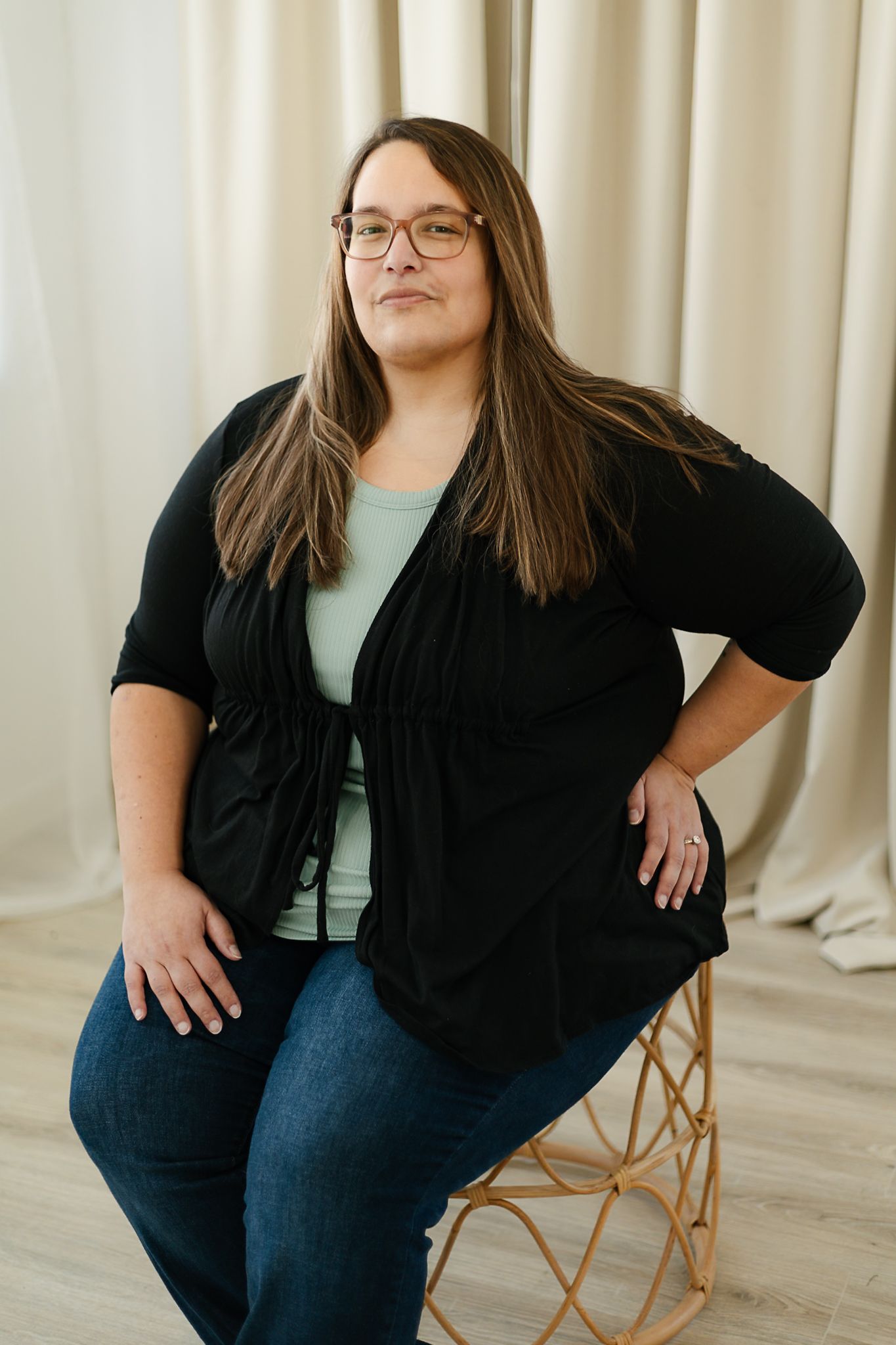 A woman in a black cardigan and mint green top smiles warmly while posing against a cream-colored curtain backdrop indoor studio, elkton SD