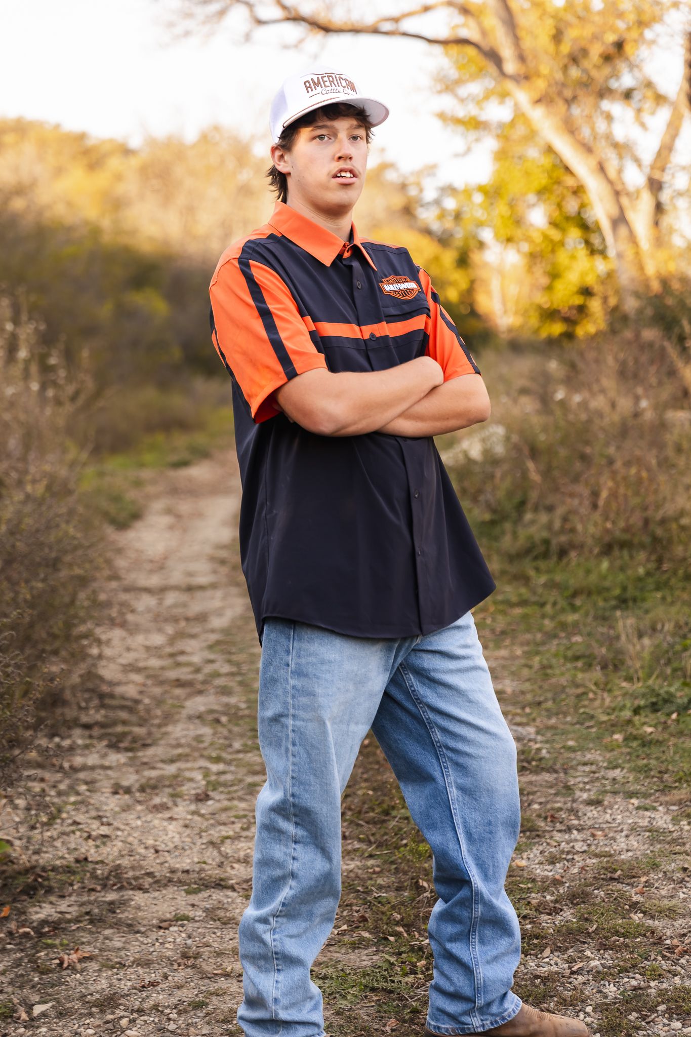 Senior in blue jeans and orange harley vest stands on dirt path during golden hour in Lake Benton, MN