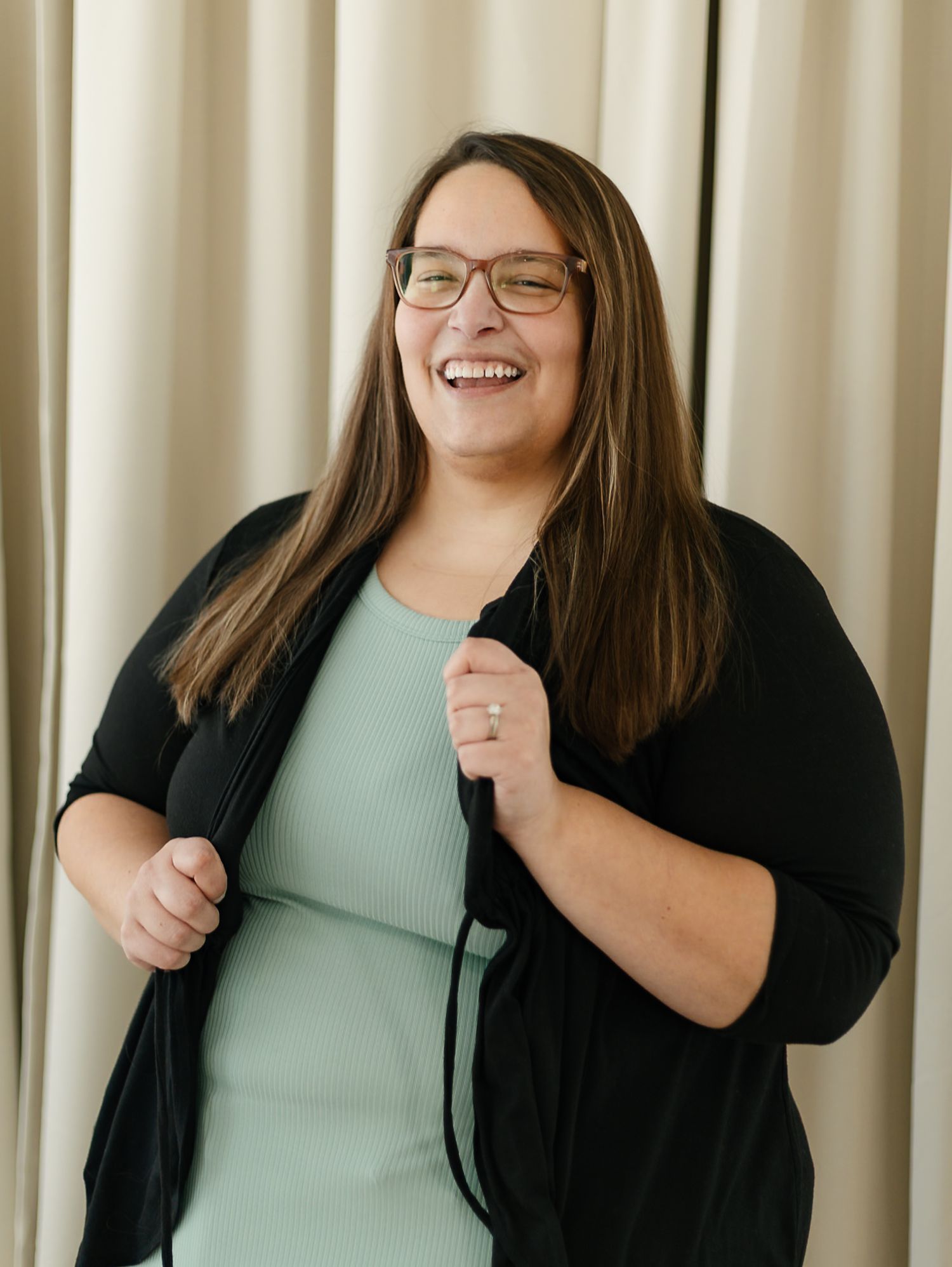A woman in a black cardigan and mint green top smiles warmly while posing against a cream-colored curtain backdrop indoor studio, elkton SD