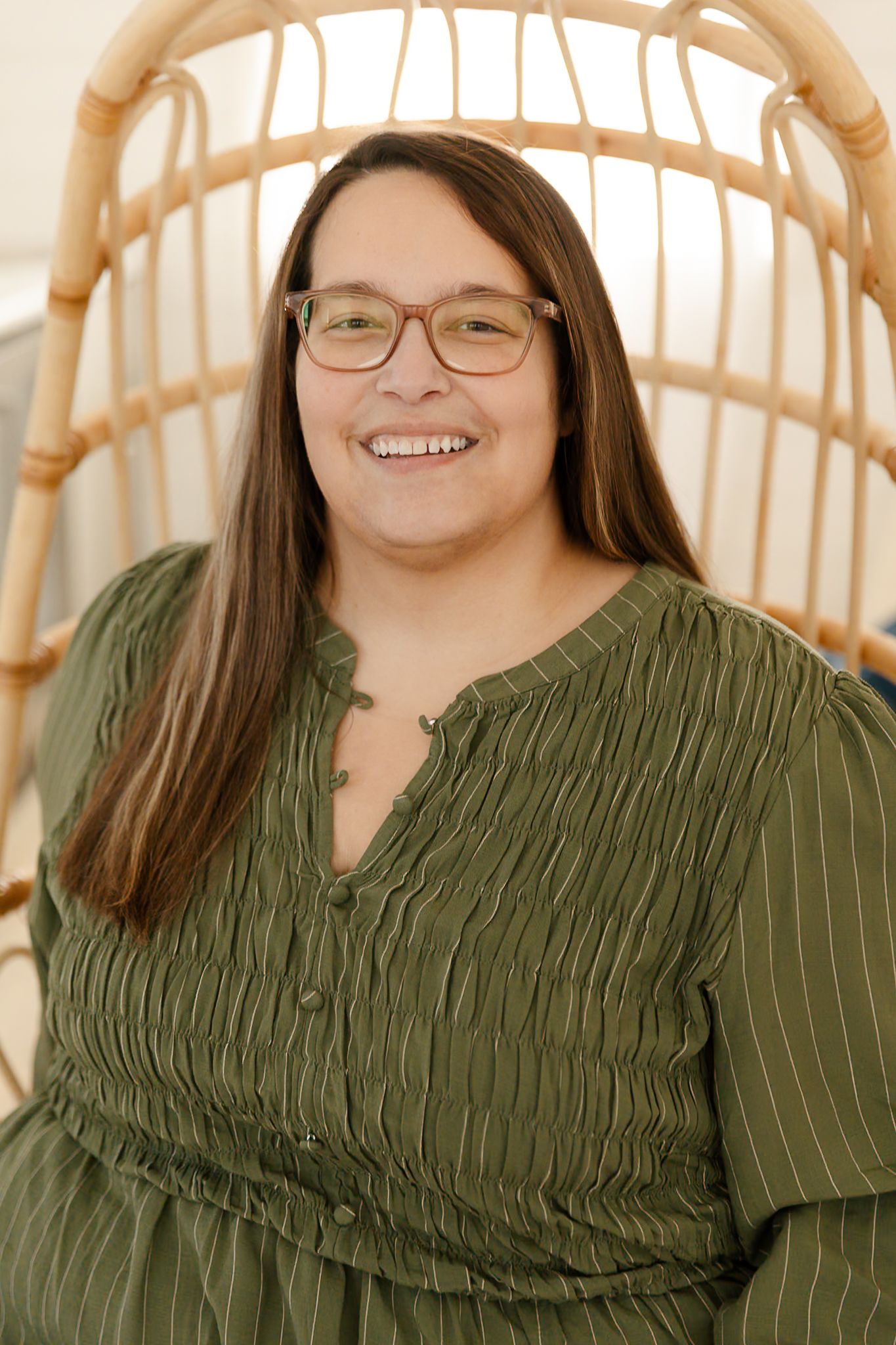 A woman in a sage green top smiles warmly while posing and sitting in a wicker egg chair indoor studio, elkton SD