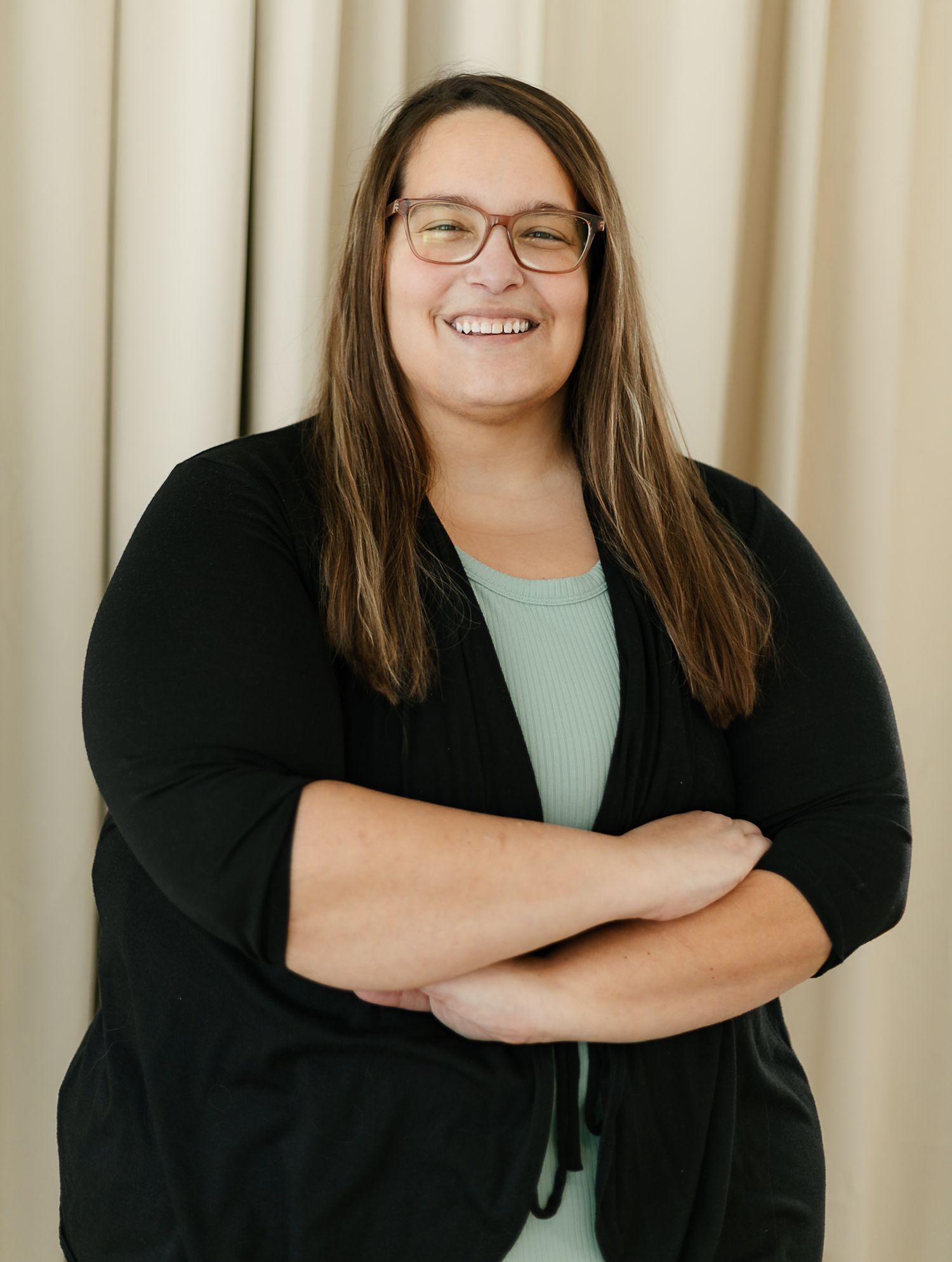 A woman in a black cardigan and mint green top smiles warmly while posing against a cream-colored curtain backdrop indoor studio, elkton SD