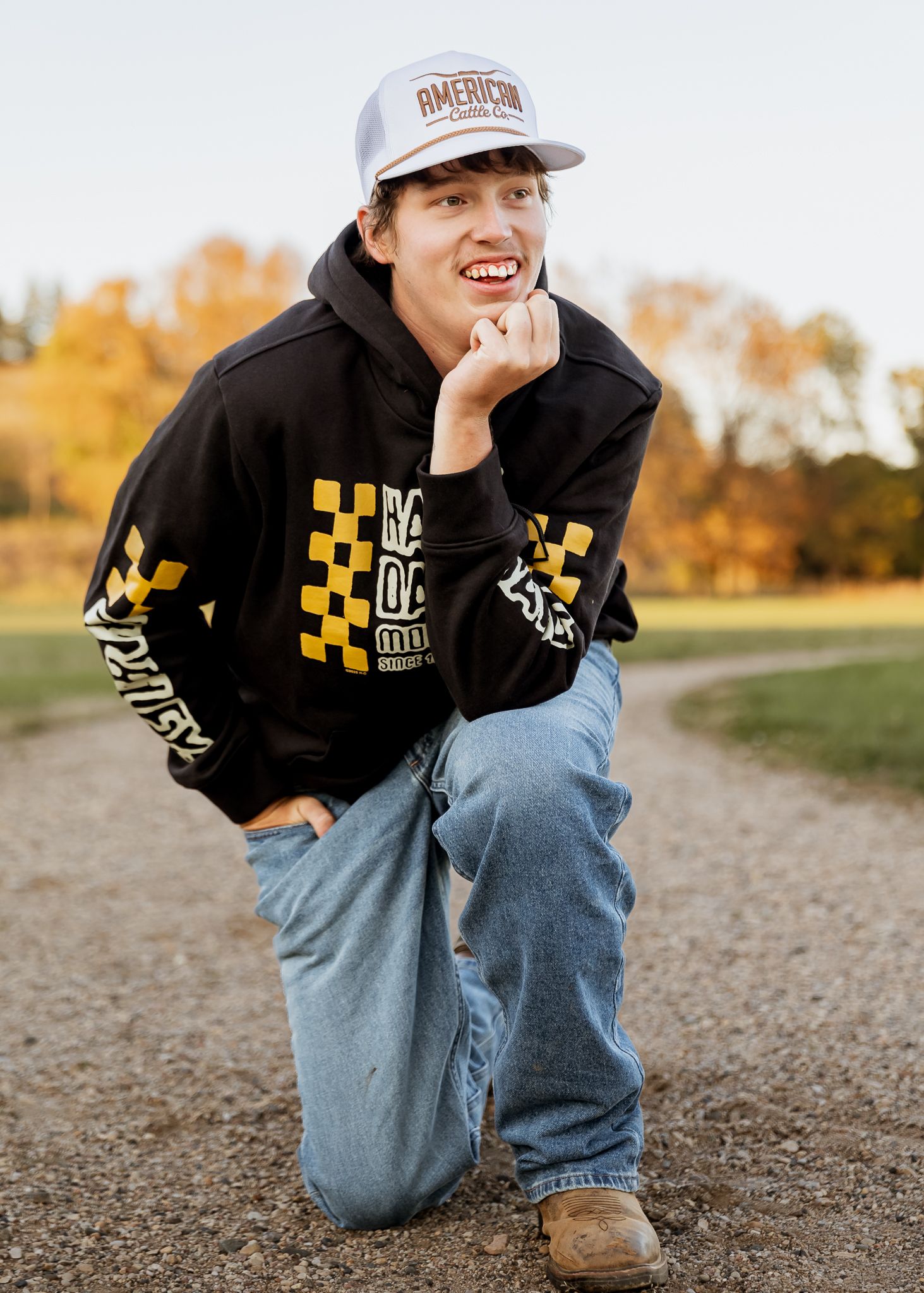 A senior in a baseball cap and black hoodie crouches on a gravel path during autumn with trees in the background in Lake Benton, MN