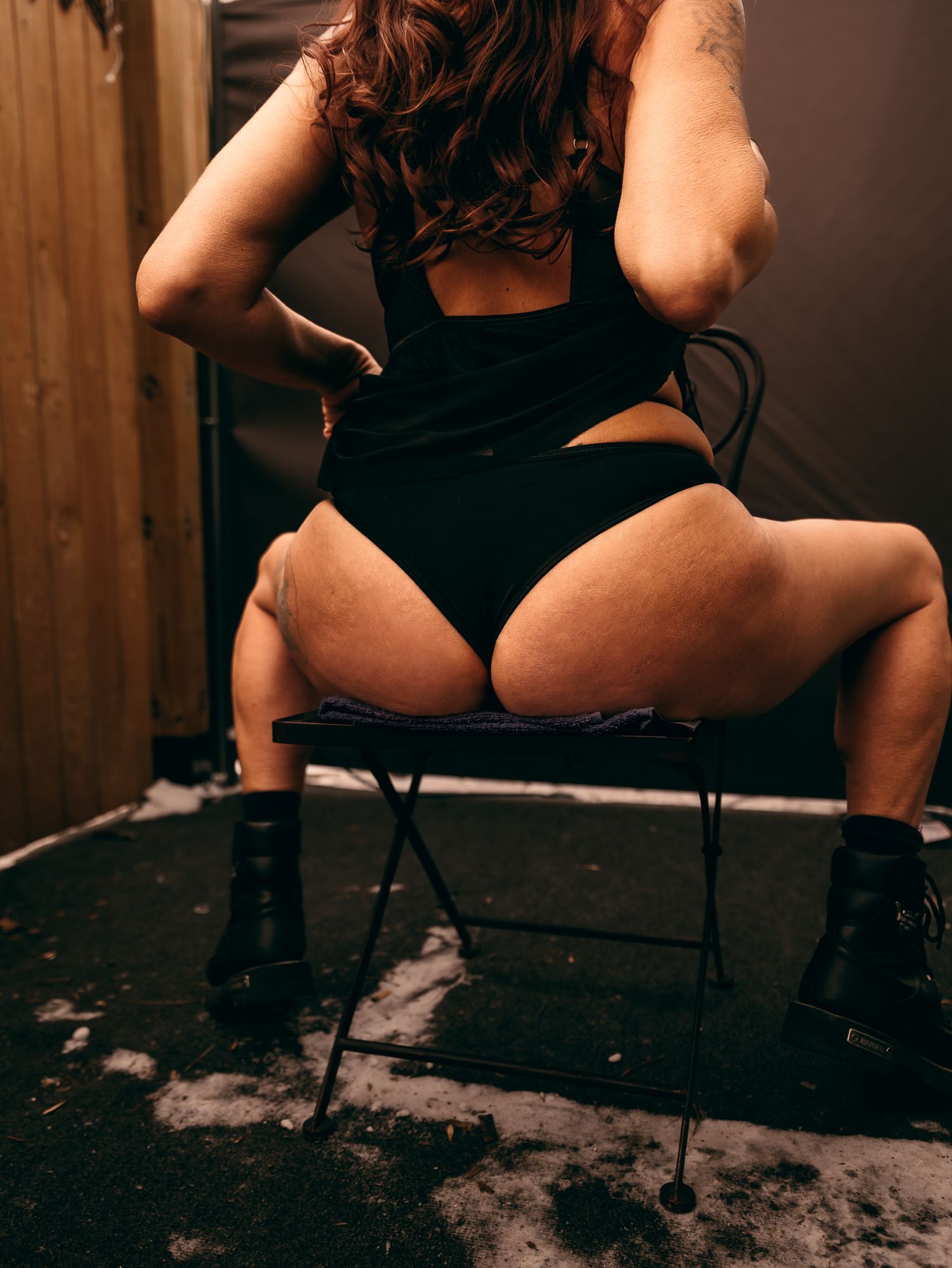 Elegant woman in black bodysuit and boots poses on a chair in a dimly lit room with dramatic lighting.