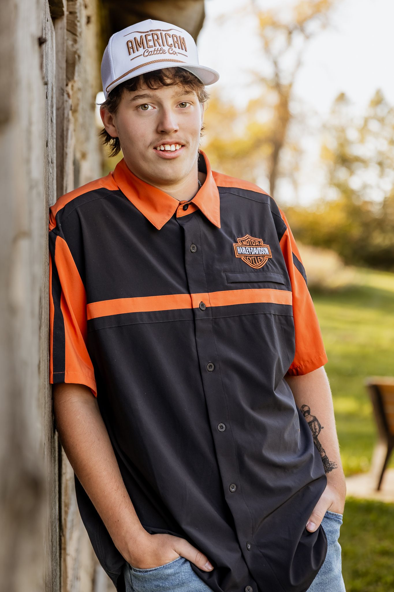 Senior wearing a Harley Davidson uniform with orange and black colors and an American baseball cap poses outdoors, lake Benton MN