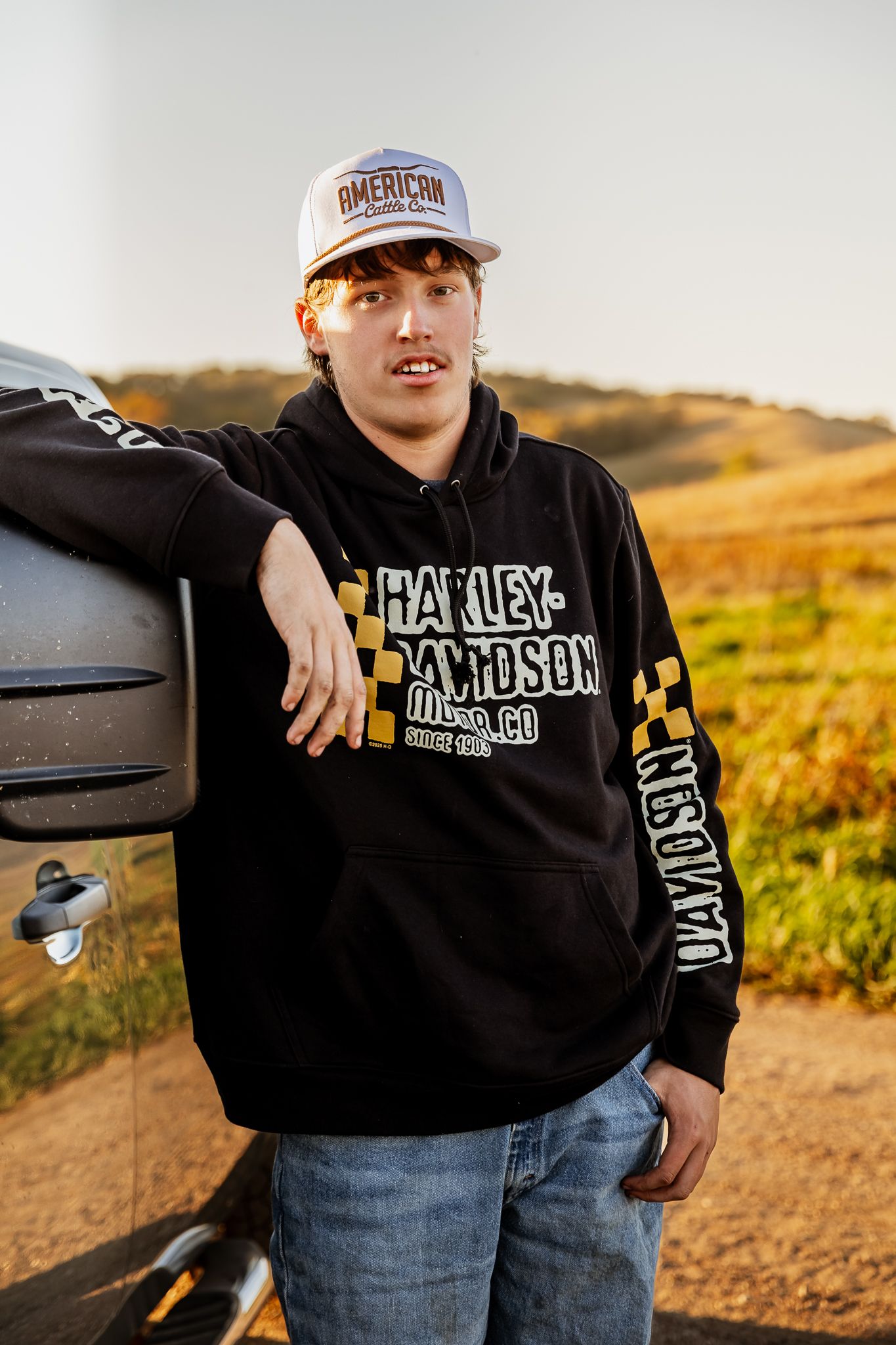 Senior wearing black Metal Mulisha hoodie and jeans poses outdoors against golden sunset landscape in Lake Benton, MN