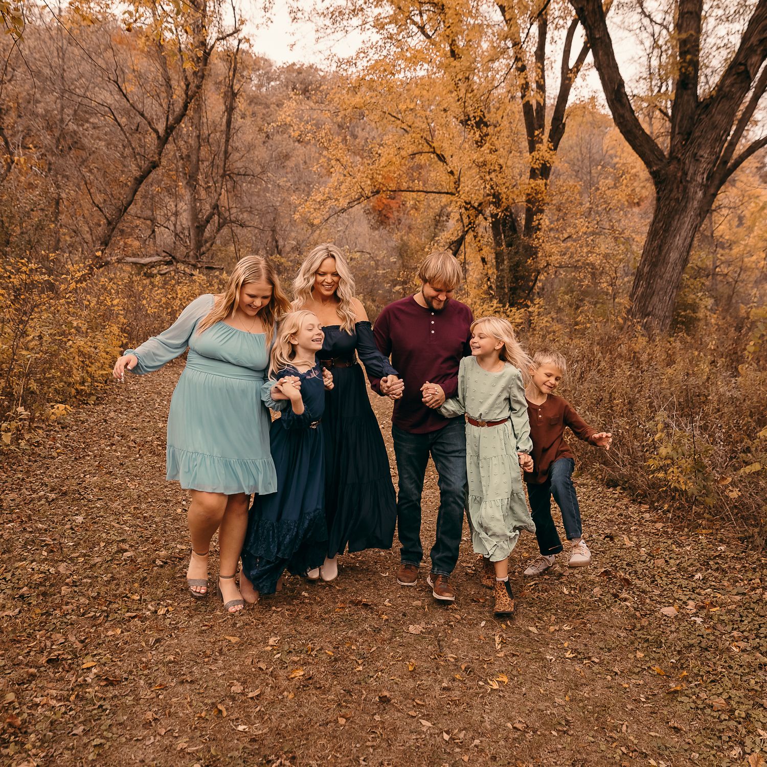 A family walking together on a leaf-covered path through autumn woods wearing coordinated blue and burgundy outfits in Lynd, MN.
