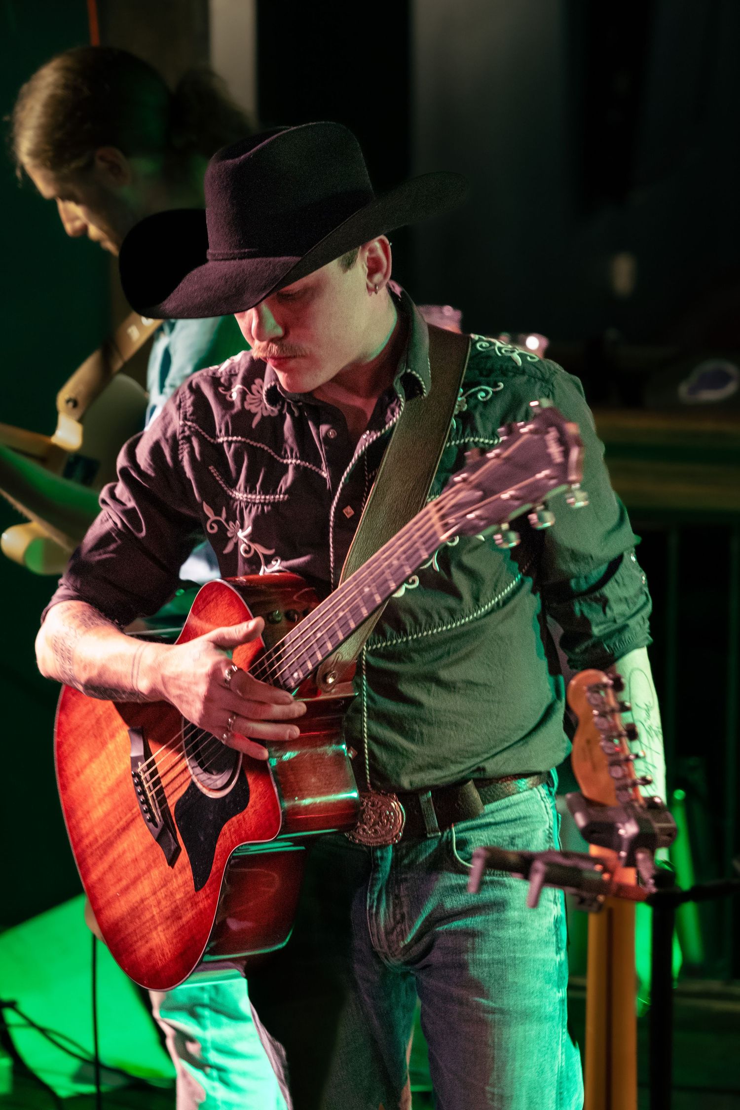 A bassist performs on stage illuminated by dramatic green and red lighting while playing a wood-finished instrument.
