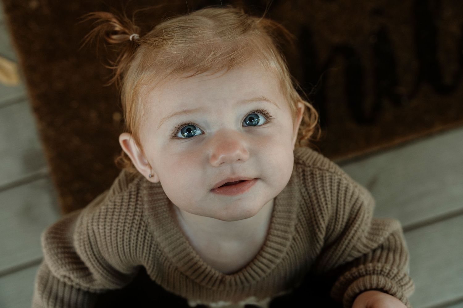 Adorable red-haired baby with blue eyes wearing a brown knit sweater looking up curiously.