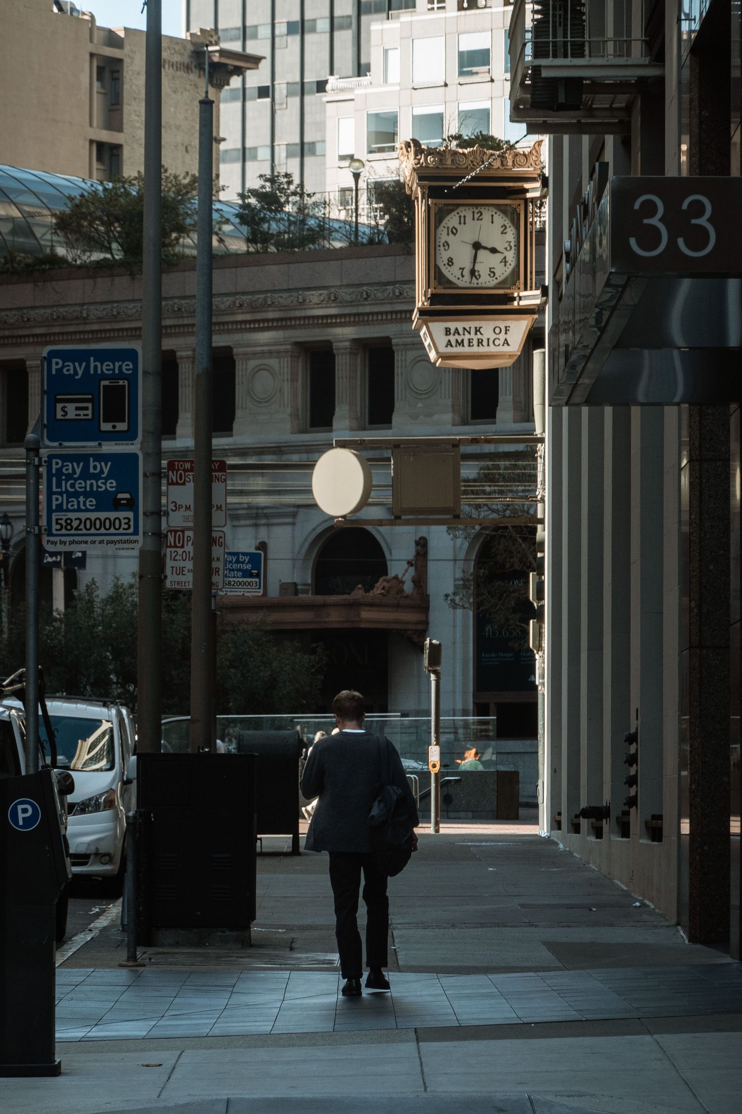 A businessman walks along a city sidewalk past a ornate Bank of America street clock on a sunny day.