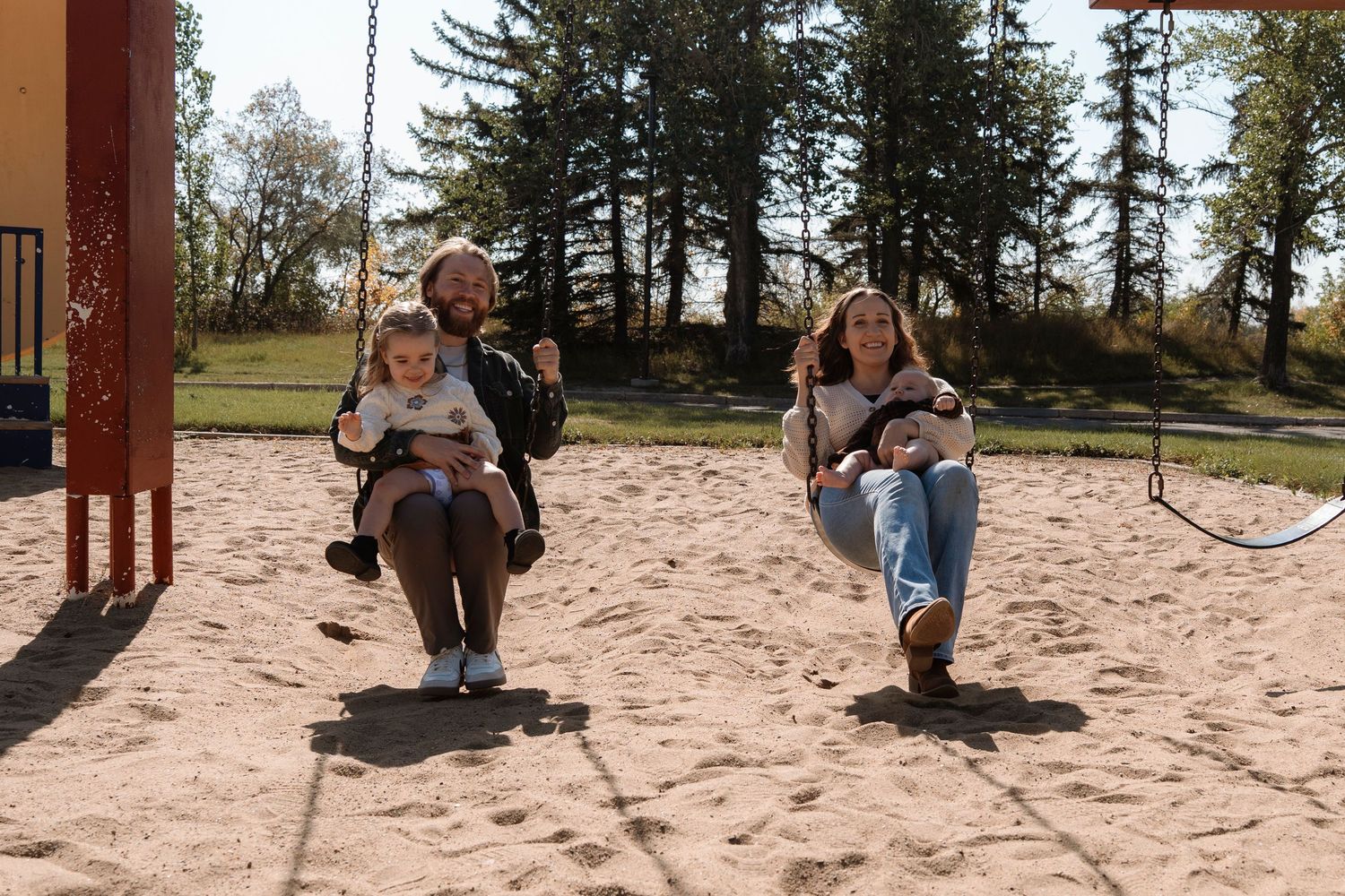 Two adults with children enjoy swinging together at a sandy playground surrounded by tall trees on a sunny day.