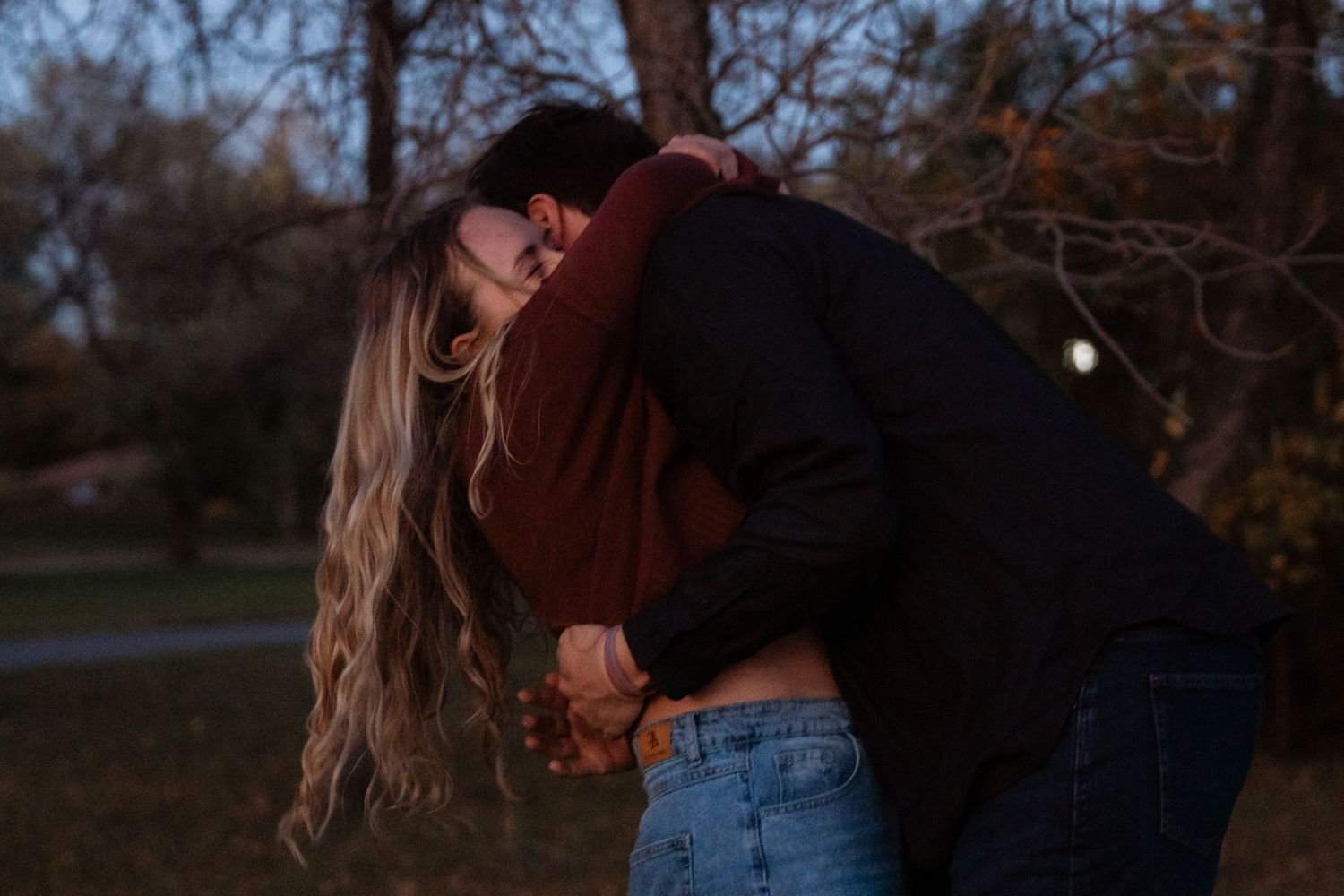 Couple sharing a romantic embrace outdoors at dusk, surrounded by autumn trees in a park setting.