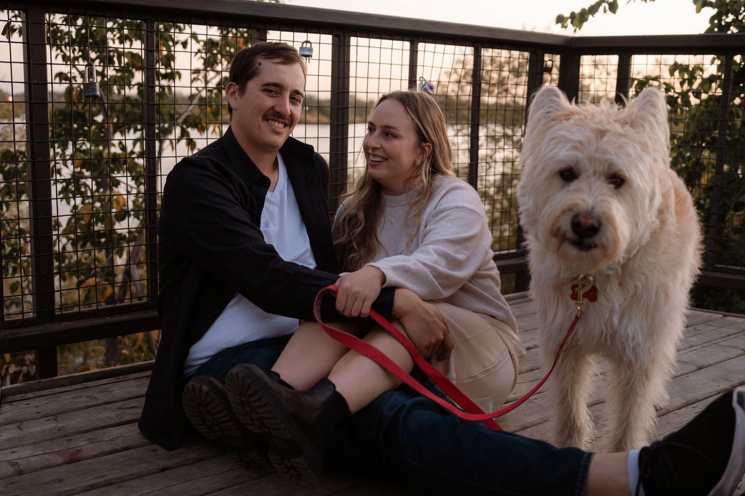 Couple sitting on wooden deck with white fluffy dog on red leash at sunset.
