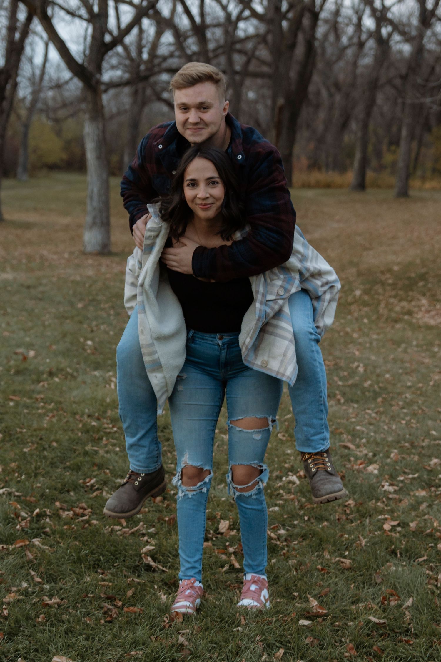 Young couple in jeans enjoying a piggyback ride in an autumn park, smiling and embracing outdoors.