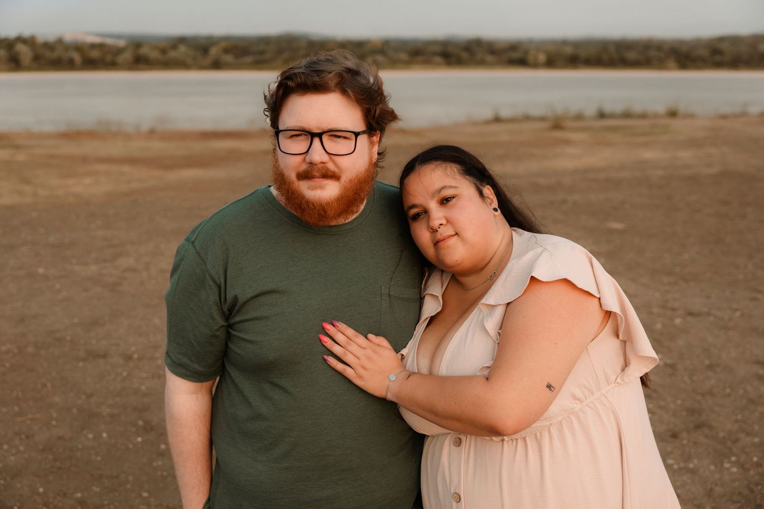 Couple posing outdoors near a lake, man in green shirt with red beard, woman in pink dress leaning on him.