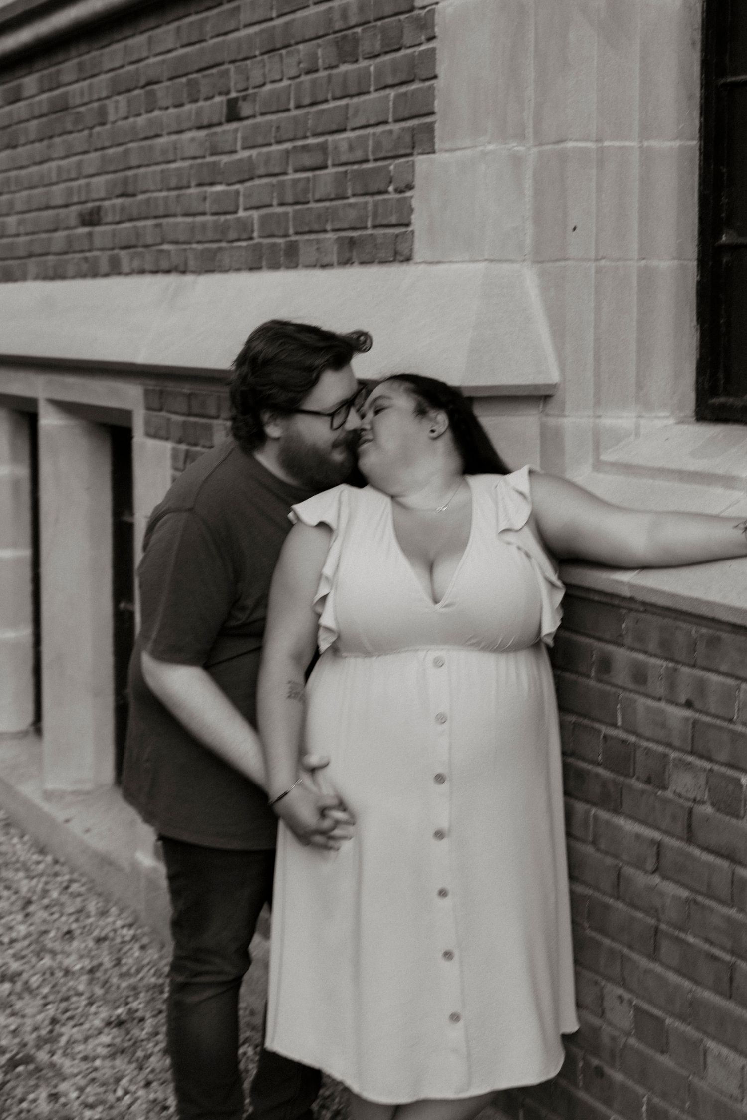 Black and white photo of a couple sharing a romantic moment against a brick wall, woman in white dress.