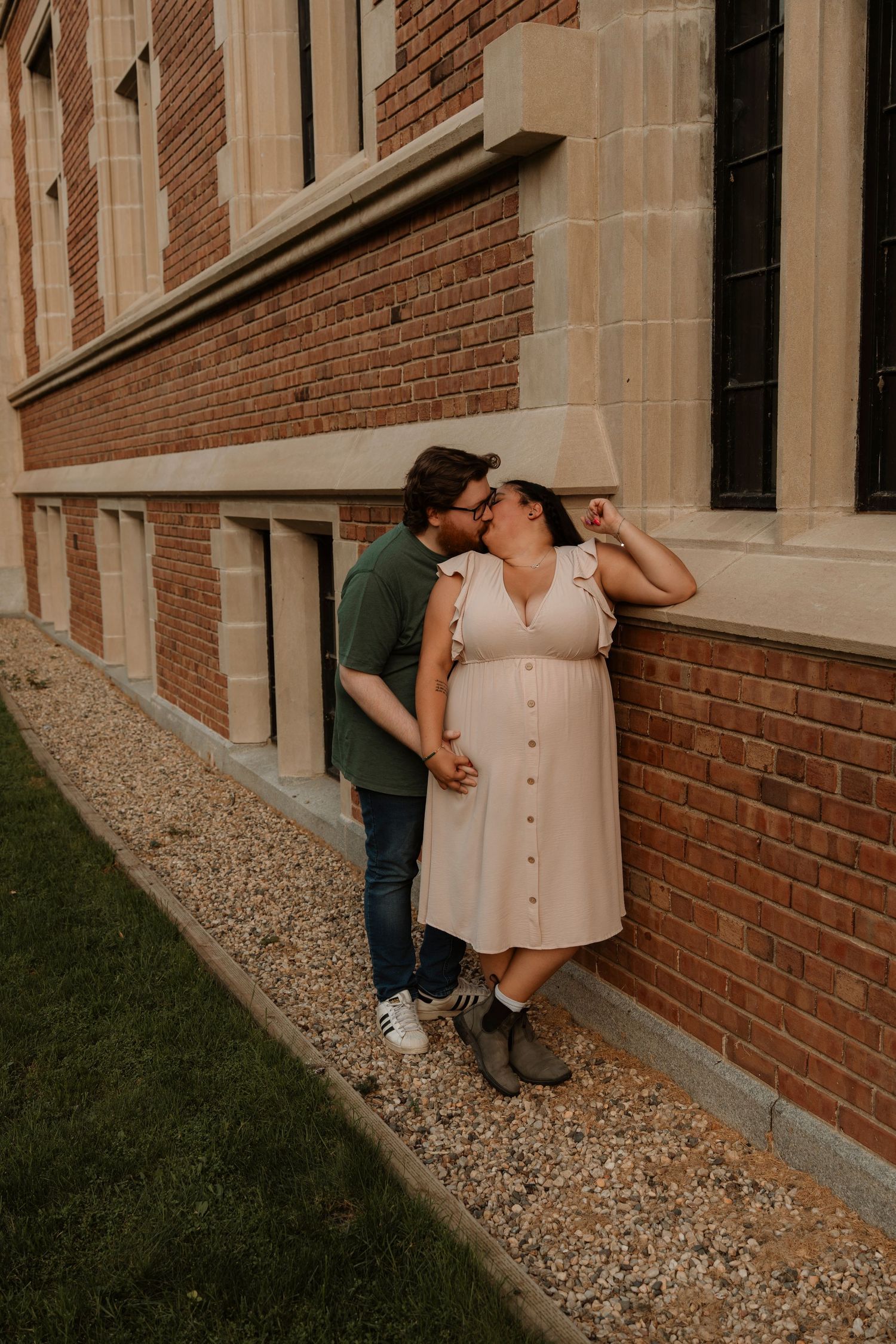Pregnant couple sharing a kiss against a brick building wall, woman in pink dress holding her baby bump outdoors.