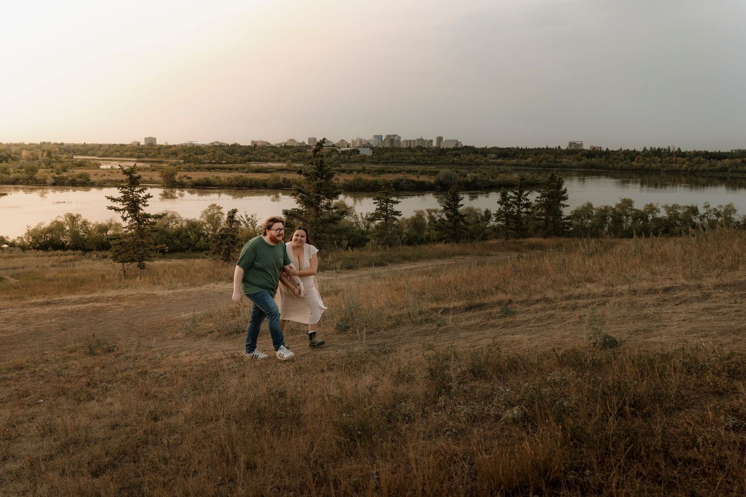 Couple walking hand in hand through golden field at sunset with river and trees in background.