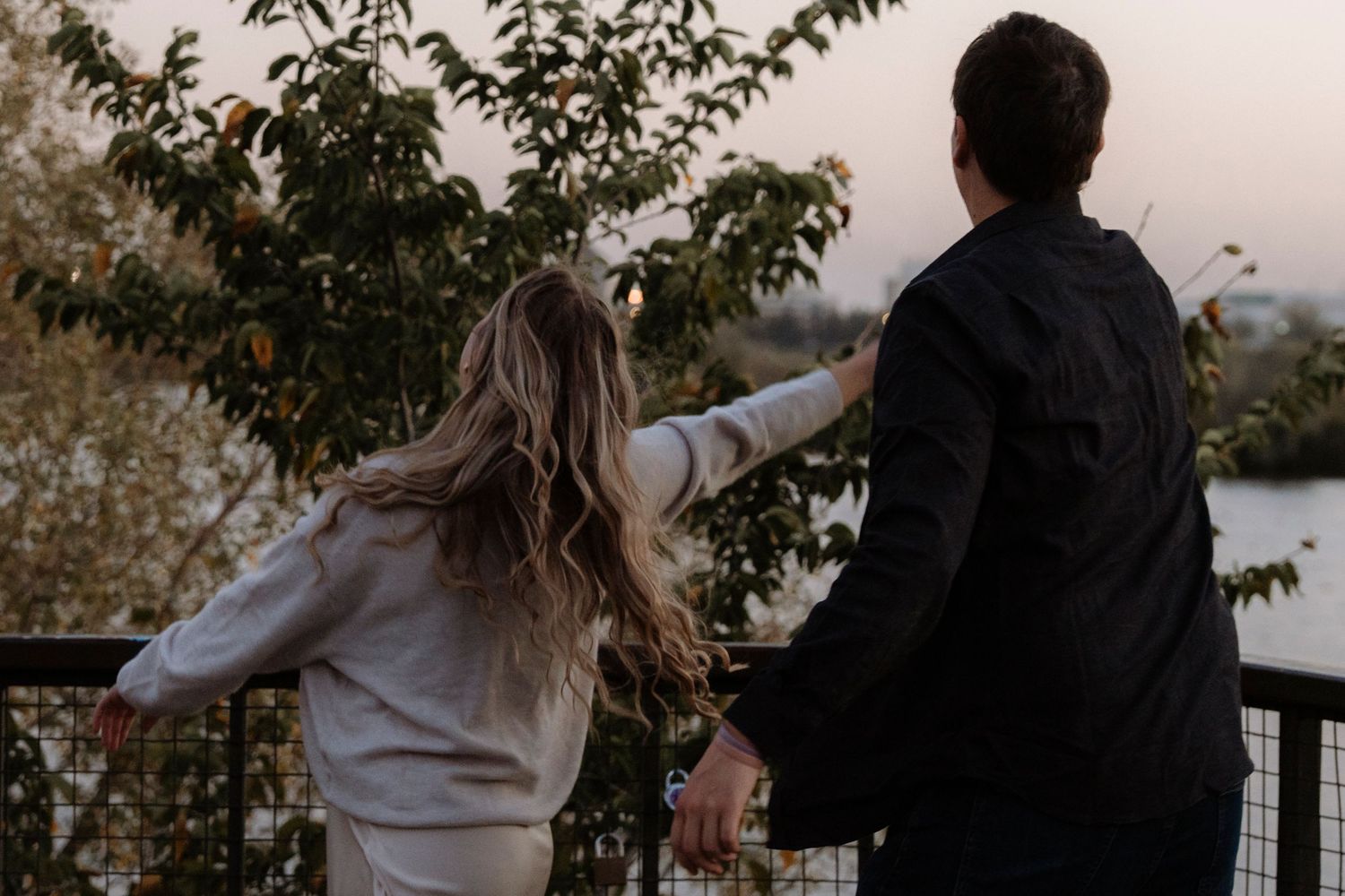 Couple holding hands at a scenic overlook at dusk, surrounded by lush trees and a tranquil waterway.