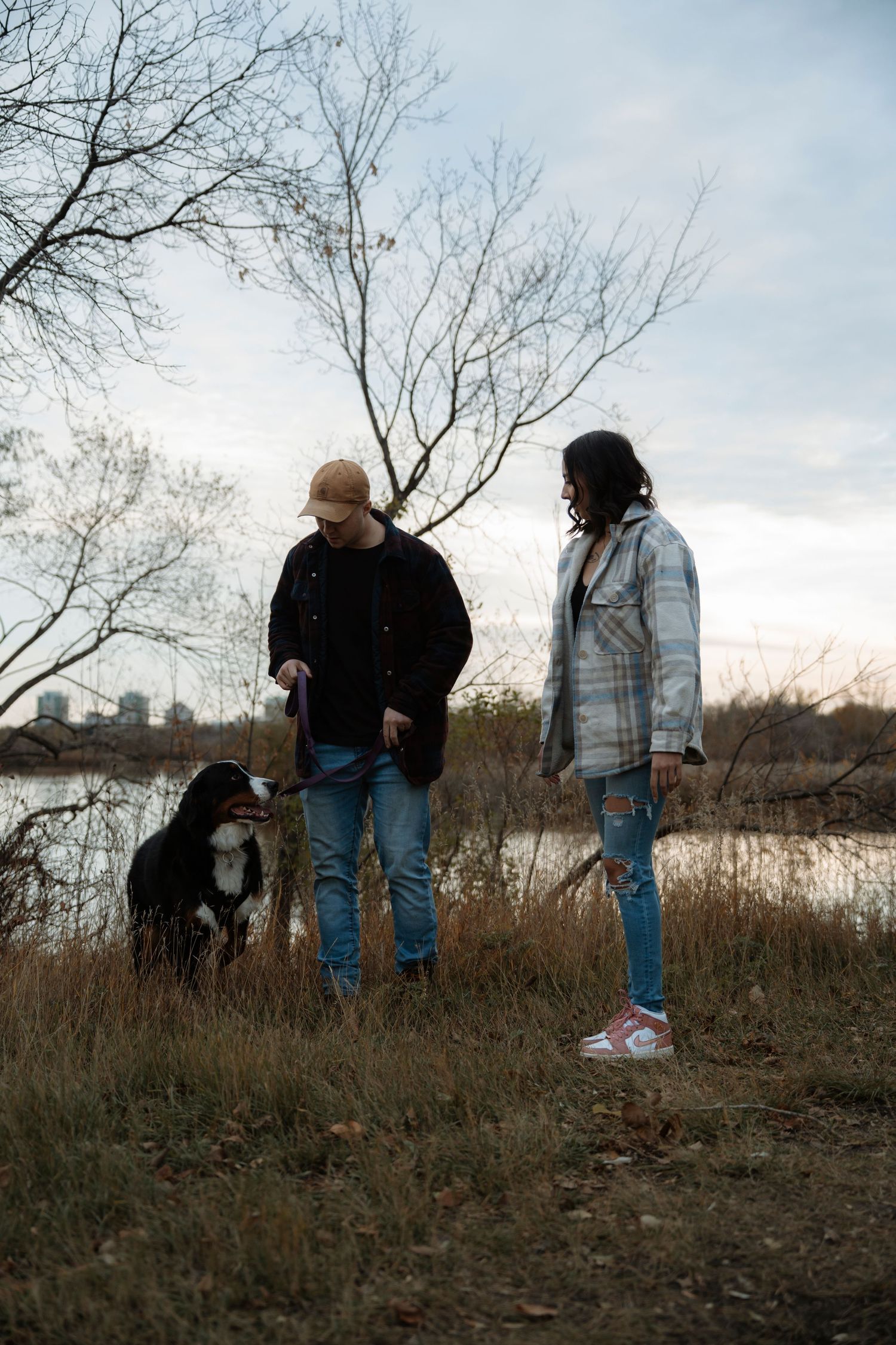Couple with Bernese Mountain Dog by a lake in autumn, surrounded by bare trees and dry grass at dusk.