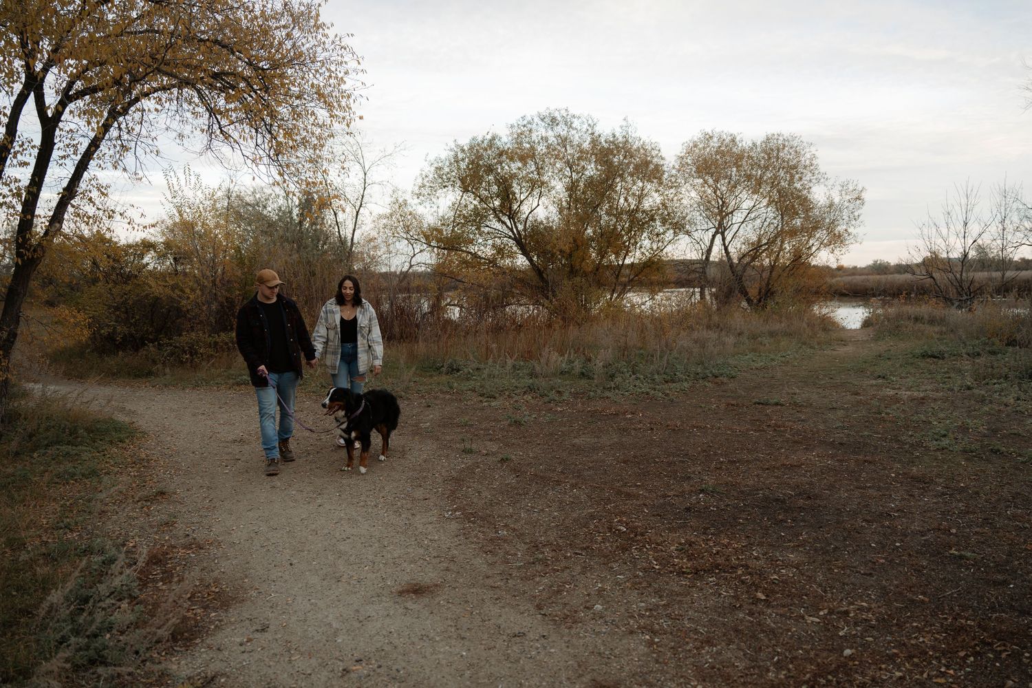Couple walking black dog on dirt path near river with autumn trees in background.