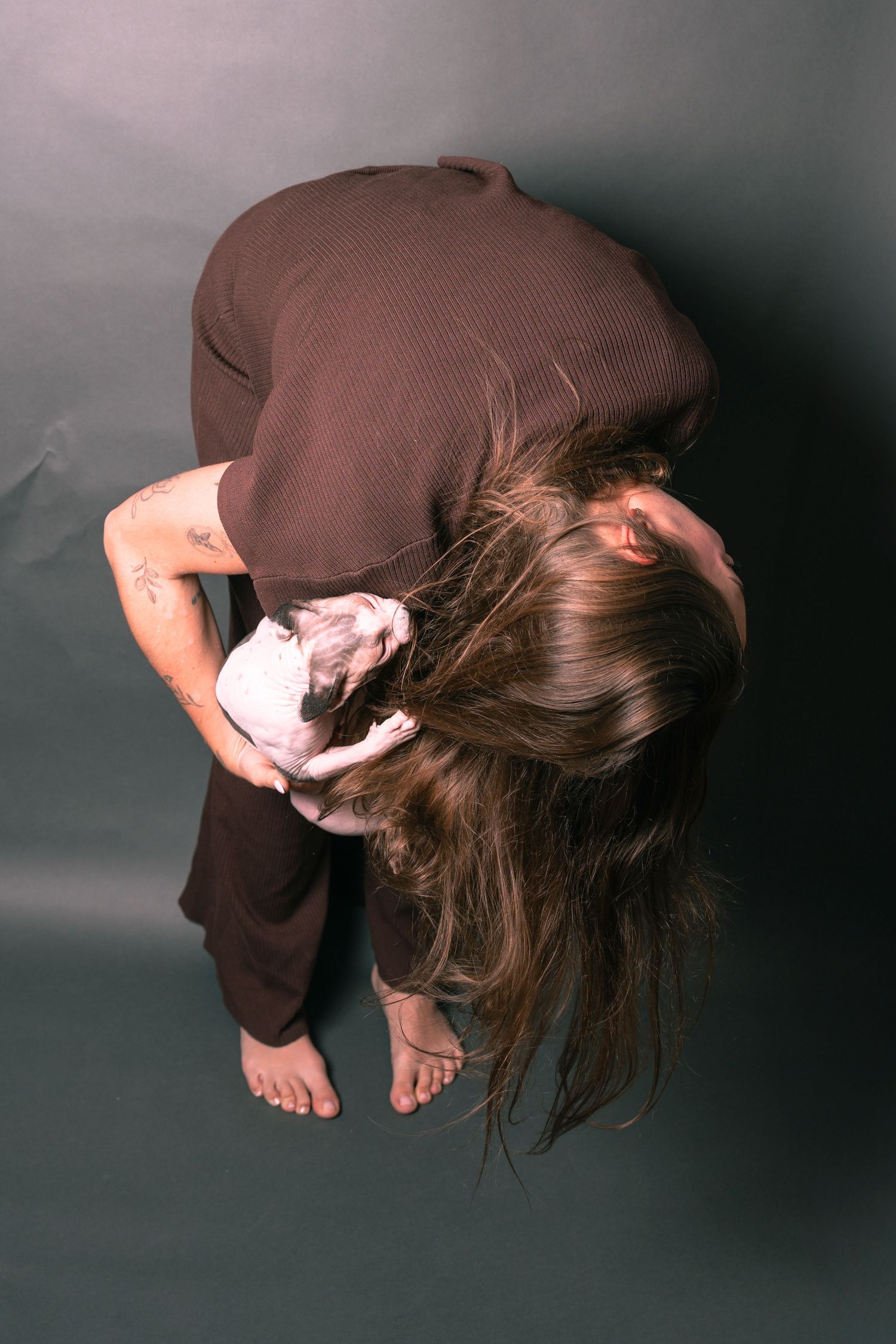 Overhead view of a barefoot woman in brown outfit bending forward with long hair falling down, holding a small hairless cat.